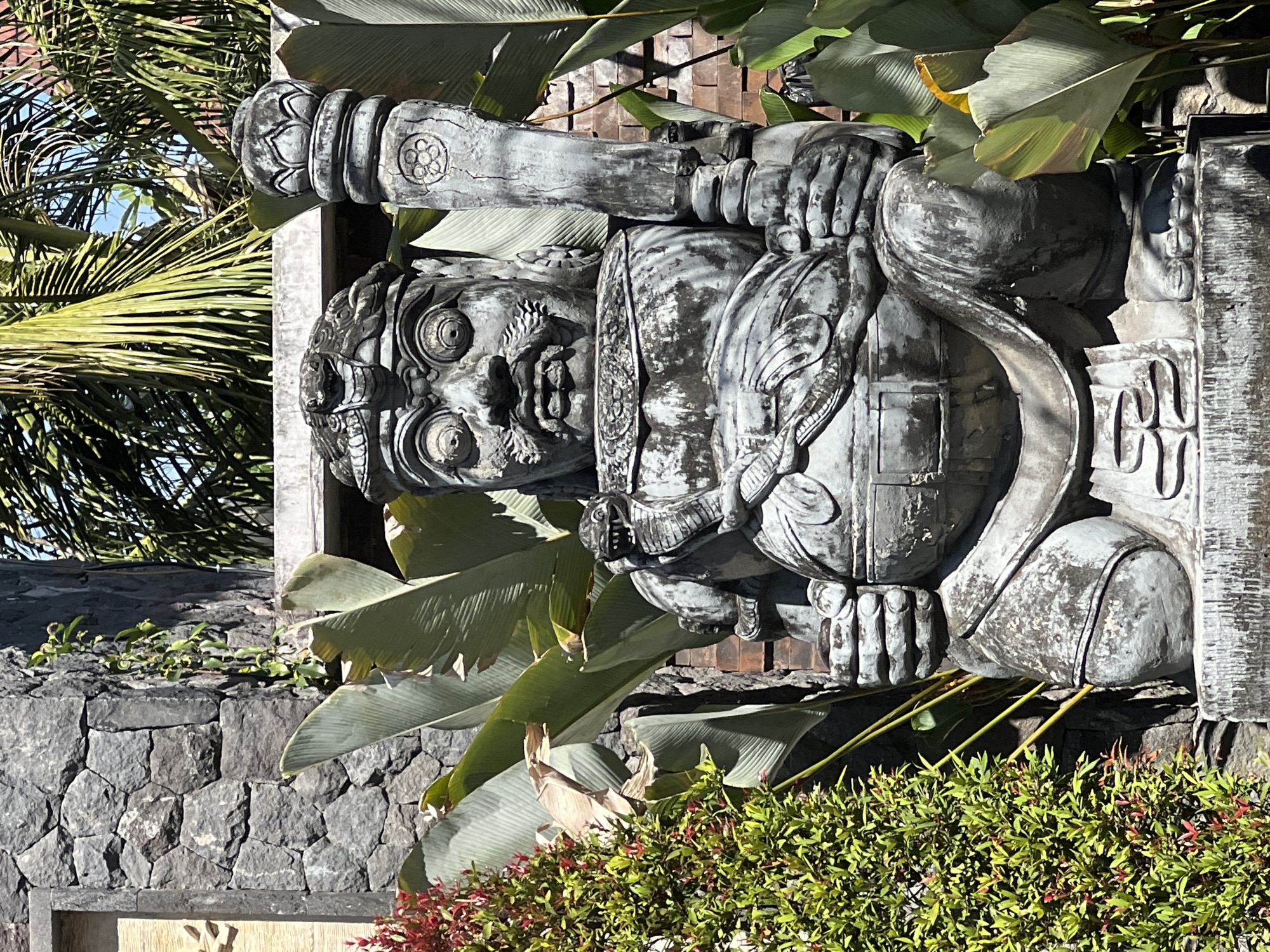 An outdoor, eye-level medium shot captures a weathered, gray stone statue of a traditional Balinese guardian figure in Sukawati, Indonesia. The statue is seated and depicts a fierce face with wide, bulging eyes, a prominent mustache, and an open mouth revealing teeth. It wears an ornate headdress and is adorned with a snake coiled around its chest and shoulder. The guardian holds a large club-like object in its right hand and its left arm rests on its lap. The statue is positioned amidst lush green foliage, including large-leafed tropical plants and a bush with red and green leaves on the left. Behind the statue, a rough stone wall and a brick wall are visible, with palm fronds extending from the top. The lighting suggests a bright, sunny day.