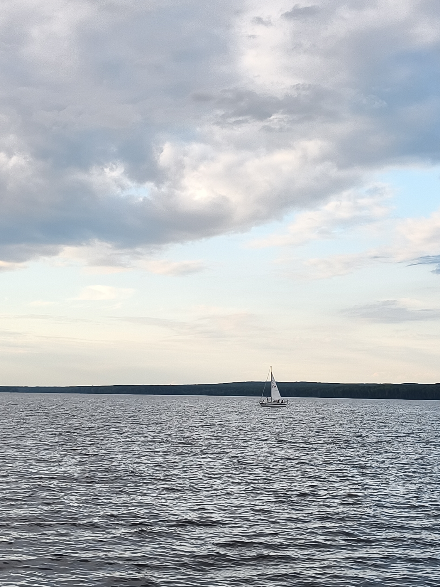 The image shows a wide, calm body of water, likely a reservoir or large lake, with gentle ripples on the surface. A solitary sailboat with a white sail is visible in the mid-ground, sailing from left to right. The boat appears to have at least two people onboard. In the background, a dense, dark green line of trees forms the horizon, suggesting a forested shoreline. Above the water and trees, the sky is filled with a mix of soft grey and white clouds, with some lighter blue patches visible. The overall scene conveys a sense of tranquility and open space. While the exact location within Chaikovsky, Russia is not definitively identifiable from the image alone, this type of expansive water body with forested shores is characteristic of areas around reservoirs or lakes near the city, such as the Kama River reservoir.