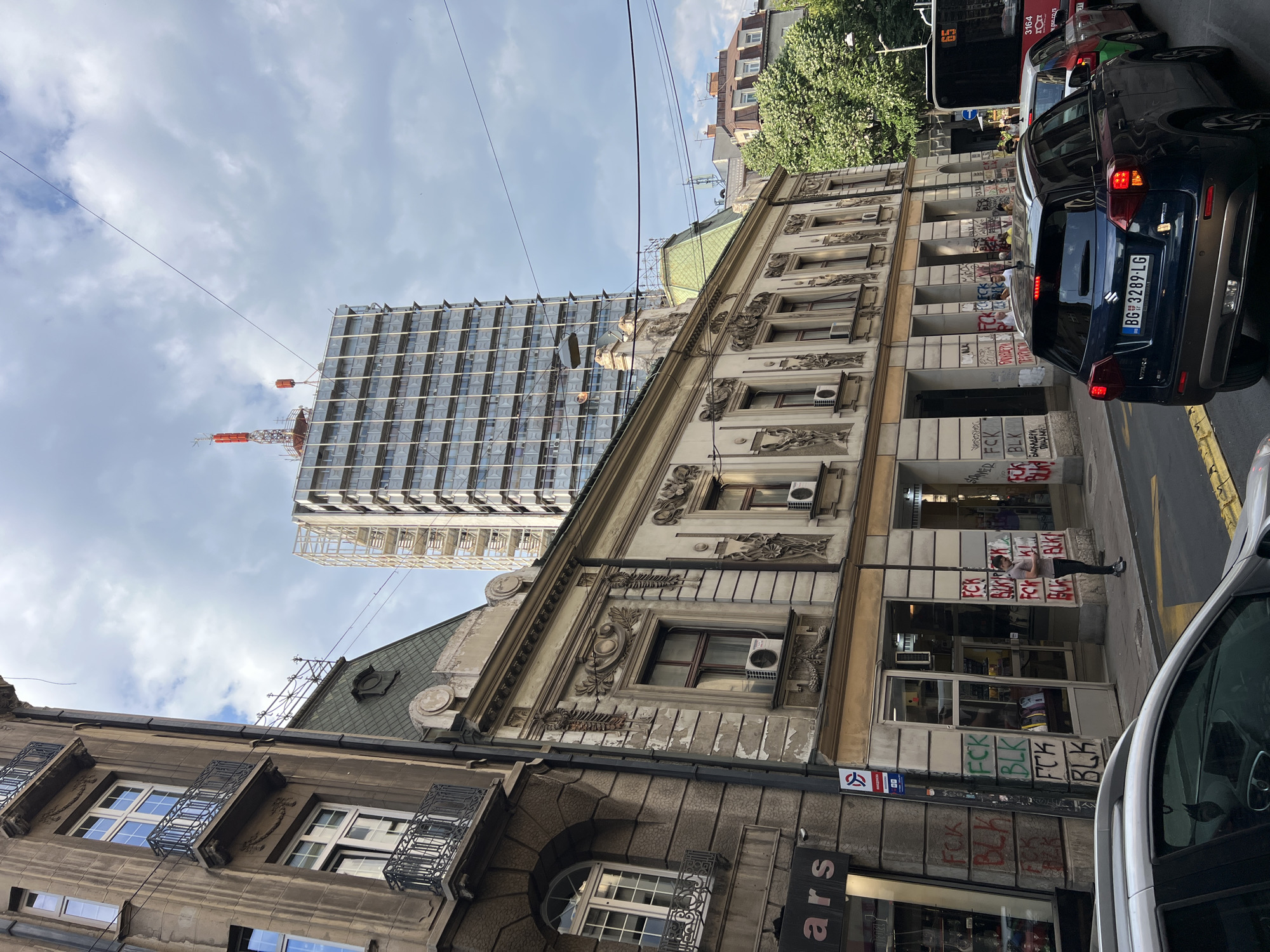 A street scene in Belgrade, Serbia, features buildings with varied architectural styles. A modern, partially constructed skyscraper with a grid facade rises in the background. An older building with ornate detailing and a green roof is adjacent to the skyscraper. Graffiti is visible on the lower portion of the older building. Several cars are parked along the street, including a dark blue SUV with license plate BG 3289-LG. The sky is overcast with clouds. Overhead wires are visible. A person is standing near the building with graffiti. A shop called "ars" is visible on a sign.