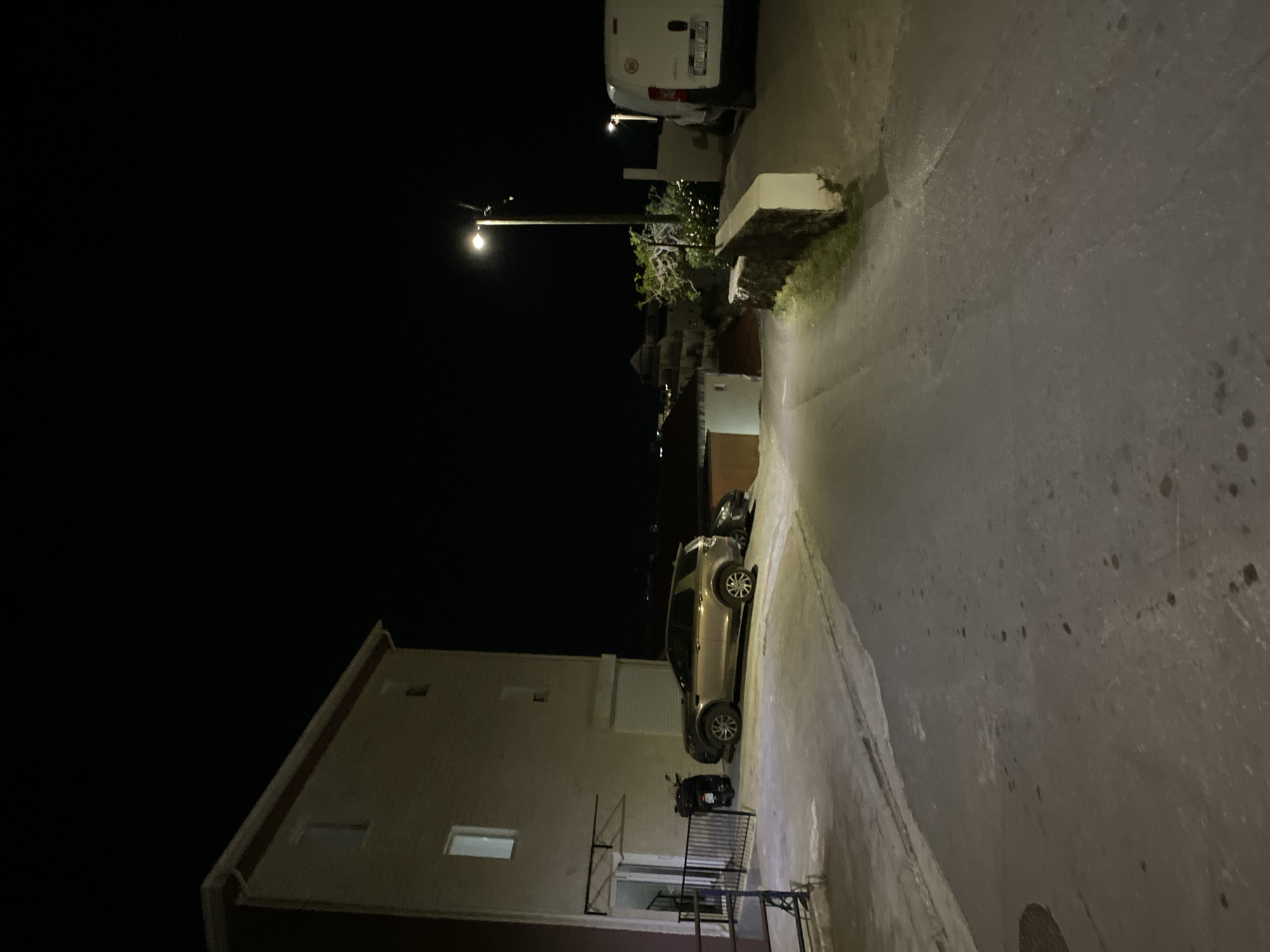 The image depicts a narrow street in Budva, Montenegro, at night. A light-colored building, possibly an apartment complex, stands on the left side of the street. Parked along the right side of the street is a tan-colored SUV and a motorcycle. In the background, a white van is parked further up the street, illuminated by a street light. The scene is dimly lit by the street light.