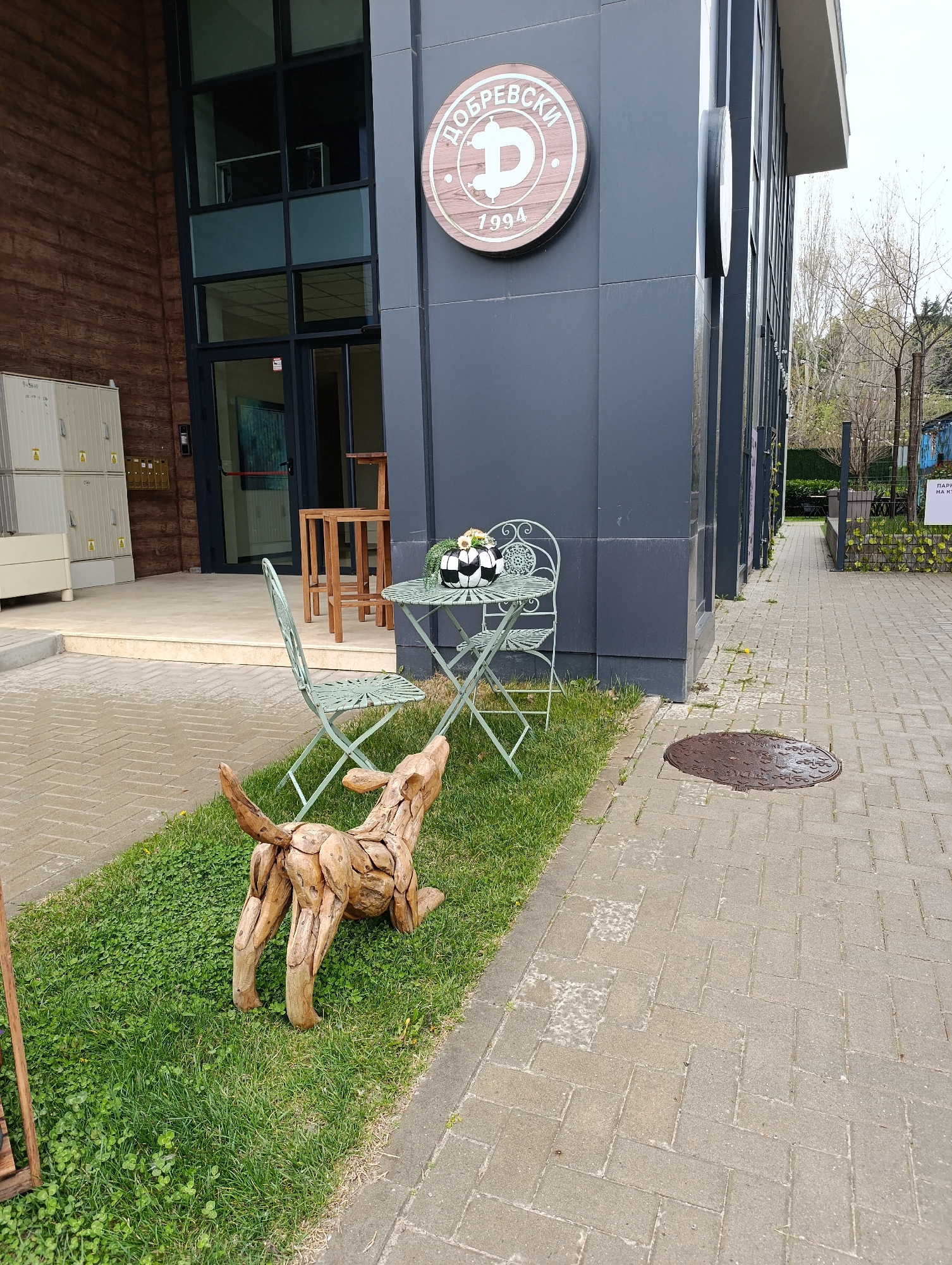 The image shows an outdoor scene in Sofia, Bulgaria. A wooden sculpture of a dog is positioned on a patch of grass next to a paved walkway. Behind the sculpture is a small, light green metal table and chair set with three objects placed on the table. Behind the table and chairs is a modern building with a sign reading "ДОБРЕВСКИ" with a "D" logo and the year 1994. The building is primarily clad in dark gray panels, and has large windows and a brown wood façade on the left side. In the background, a walkway leads into a green area with a partially visible sign in Cyrillic. The weather appears overcast.