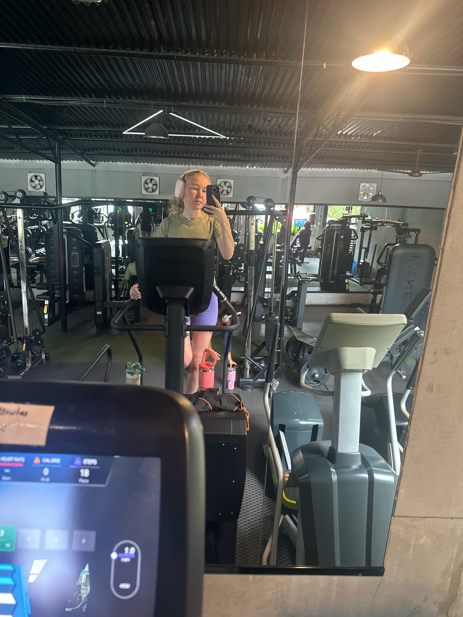 A woman with blonde hair, wearing light olive green t-shirt and purple shorts, is taking a mirror selfie in a gym. She is wearing pink headphones and holding her phone in her right hand. She is standing in front of a treadmill, with her left hand on the treadmill's handle. In the background, there are various gym machines, including other treadmills and weightlifting equipment. The gym appears to be indoors with a corrugated metal ceiling and exposed lighting fixtures. The overall scene suggests a casual workout session. The image was taken in the city of Lalitpur, Nepal.