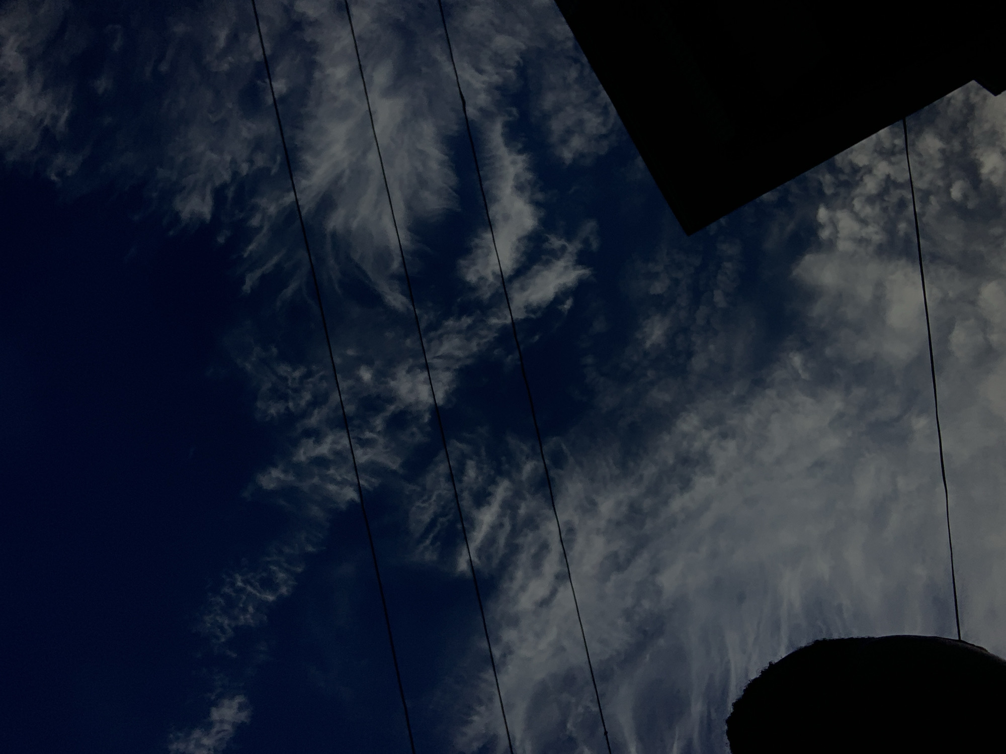A low-angle shot captures a dark night sky in Nkpor, Nigeria, filled with wispy white clouds. Four parallel power lines stretch across the frame, intersecting the celestial expanse. In the bottom left corner, the silhouetted curve of what appears to be a large, rounded object, possibly a head or a dome, is visible. To the right, the sharp, dark silhouette of a building corner juts into the frame from the bottom right. The overall impression is one of a stark, urban nightscape dominated by the dramatic sky and man-made structures.