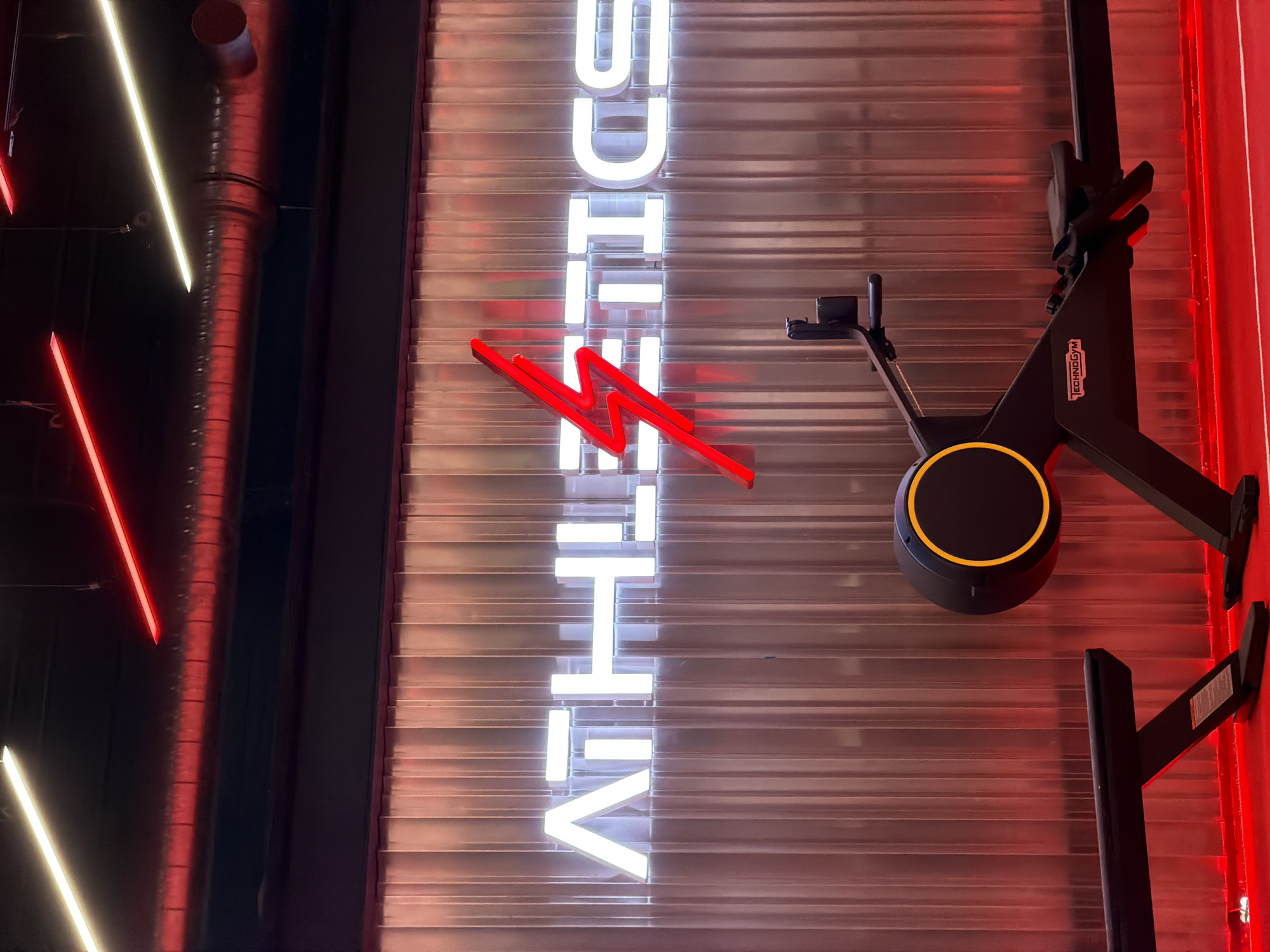 This is a medium-shot, eye-level, vertical, indoor photograph of a rowing machine in a gym. The gym has a modern aesthetic with red and white neon lighting. The word "ATHLETICS" is spelled out in large, white neon block letters on a corrugated metal wall. A red lightning bolt symbol is placed between the letters "H" and "E". The rowing machine, branded "TECHNOGYM", is black with a yellow circular accent on its flywheel. The floor is a bright red, likely a mat or painted surface. The background wall is made of vertical corrugated metal panels with a reddish hue. The ceiling has exposed conduits and two long neon light fixtures, one white and one red. The location is the Technogym showroom located at 7 Rue de Sébastopol, 67000 Strasbourg, France.