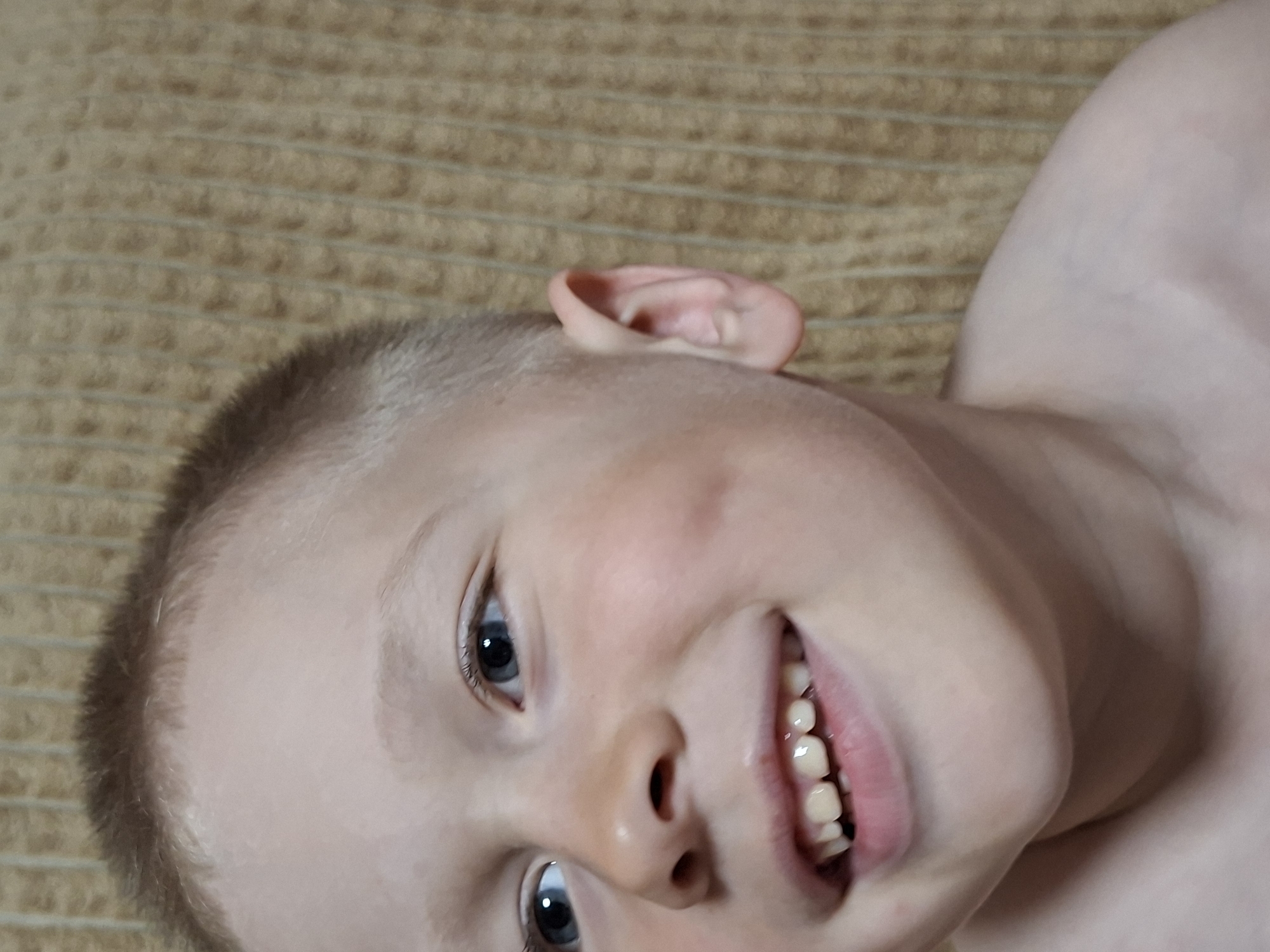 A young boy is lying on a tan textured surface, possibly a bedspread or blanket. He is fair-skinned with light reddish-brown hair cut short. The boy has bright blue eyes and is smiling, revealing his teeth. His ear is visible on the upper left side of the image. The setting appears to be indoors. No specific location information is available.