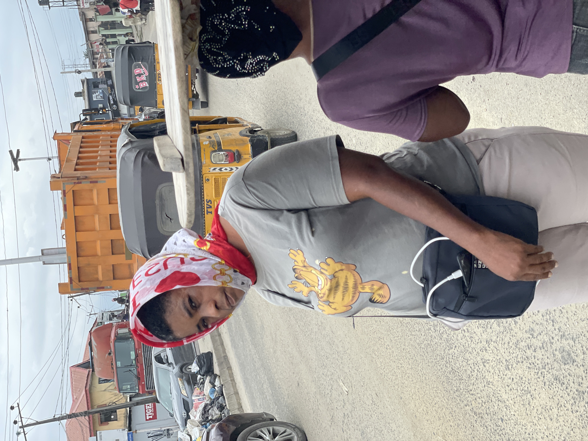 A woman wearing a grey t-shirt with a Garfield print and a red and white headscarf stands on a dusty street in Ado, Nigeria. She has a dark blue messenger bag slung across her body, with a white charging cable and a smartphone visible in an outer pocket. Behind her, traffic is heavy, with a large orange dump truck and several yellow auto-rickshaws, known as "Keke NAPEPs," on the road. The street is lined with buildings and power lines, and there are piles of trash on the side of the road. Another person, seen from behind and wearing a purple t-shirt and a black headscarf adorned with rhinestones, stands next to the woman, facing away from the camera. The overall impression is of a busy, urban environment.