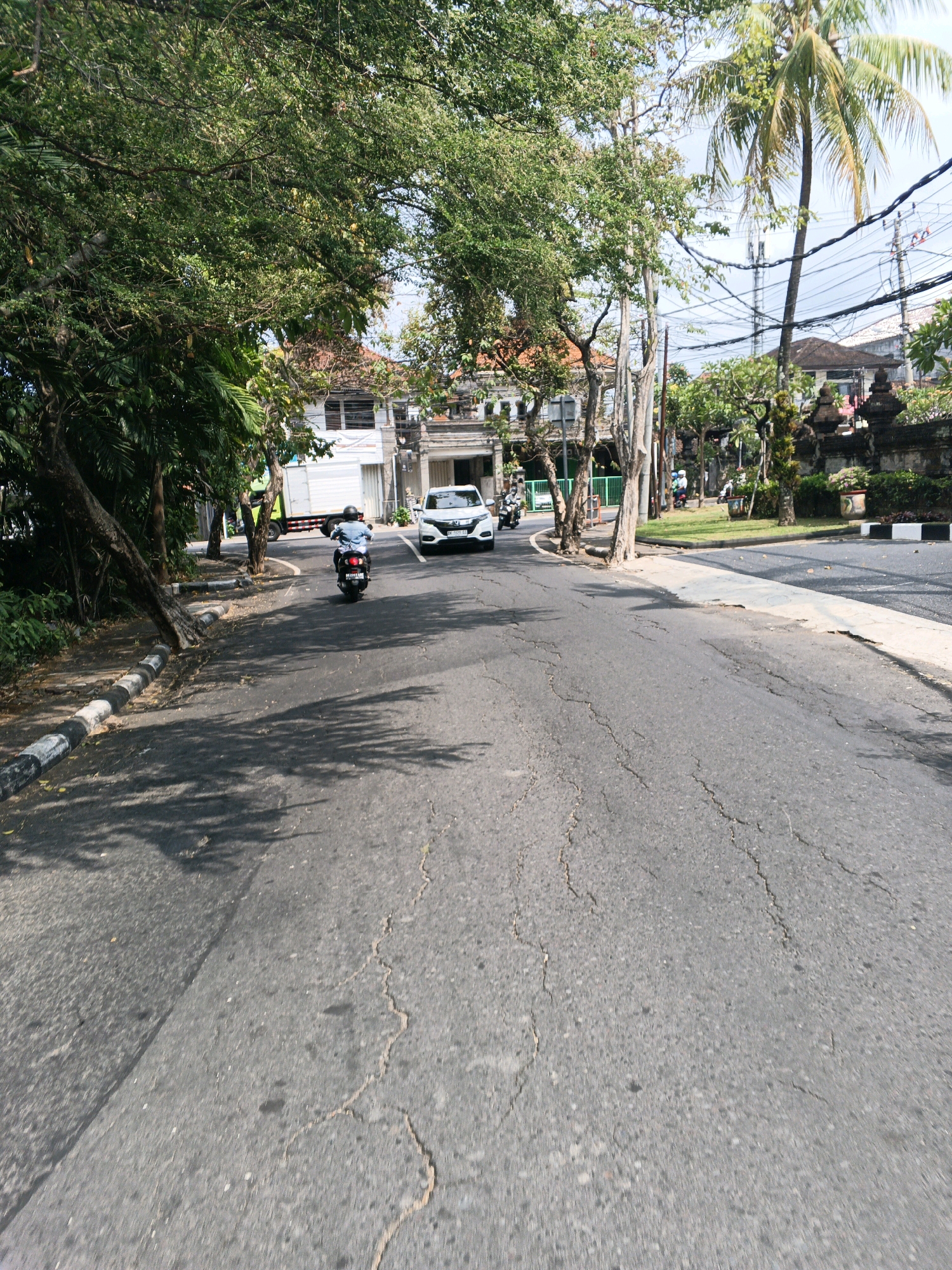 The image captures a street scene in Denpasar, Indonesia, on a sunny day. The perspective is from a low vantage point, likely a vehicle or a person on a motorcycle, looking forward along a cracked asphalt road. Lush green trees line the left side of the road, casting dappled shadows on the pavement. In the background, buildings with terracotta roofs are visible, along with palm trees, power lines, and ornate stone walls, suggesting a tropical urban environment. A white car is traveling in the same direction as the viewer on the right side of the road, and a person on a scooter is in front of the car. The overall impression is a typical street in a warm climate, with vegetation and vehicles coexisting.