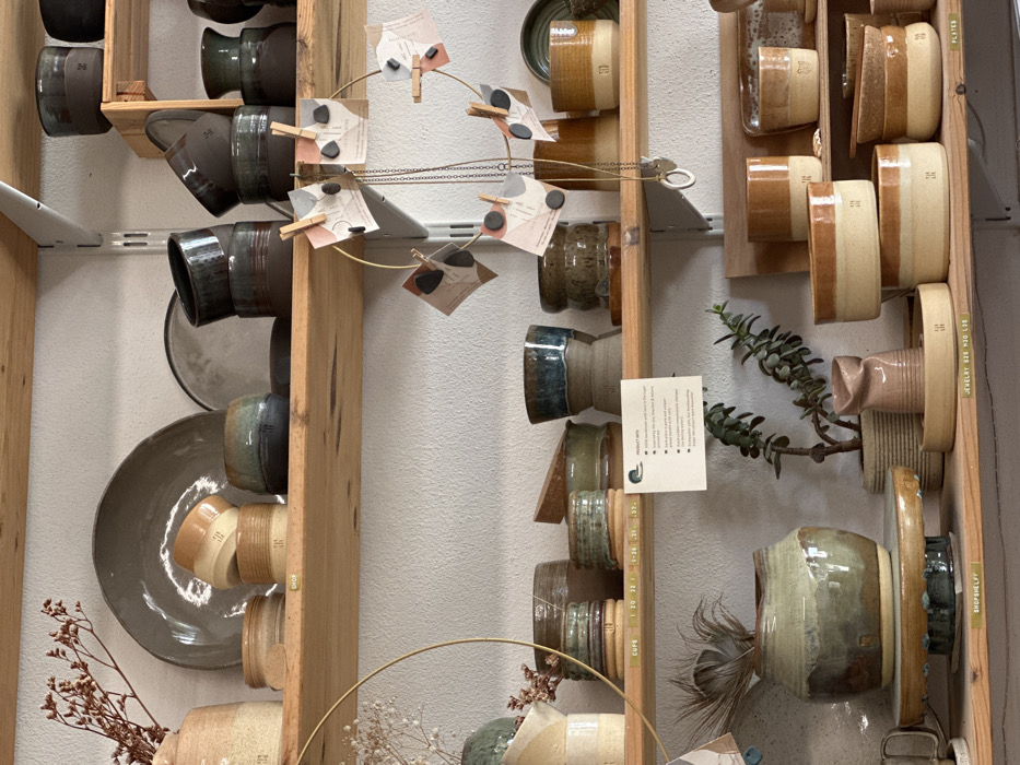 The image displays an interior retail or studio space located in Mafra, Portugal, showcasing a curated collection of handcrafted ceramic pottery arranged on three light-wood shelves against a textured, light-colored wall.
On the top shelf, from left to right, a beige ceramic vase contains dried reddish-brown botanical elements. Behind it, a large, dark grey ceramic plate leans vertically. In front, two stacked, two-toned light brown ceramic cups or bowls with darker bases are present, followed by a squat dark brown bowl featuring a greenish-blue rim. Another dark grey plate is partially visible, behind several dark brown ceramic cups and bowls with glossy dark glazes, some stacked.
The middle shelf features a ceramic pot with dried white flora on the far left. A brass-colored circular frame, adorned with small paper tags secured by mini clothespins, hangs in the foreground, from which a necklace with a ring pendant is suspended. Various ceramic cups and small bowls display diverse glazes, including striped patterns in green, brown, and cream. A white "PRODUCT INFO" card is centrally positioned. To the right, a two-toned cup with a deep blue upper glaze and a light brown base is visible, along with additional brown and dark-glazed ceramic containers, one bearing a subtle "TH" or similar logo.
The bottom shelf on the left supports a large, rounded ceramic pot with a mottled greenish-brown glaze, resting on a circular wooden stand with a turquoise rim. A single feather is placed inside the pot. A white label below the stand reads "SHOPSHELF." To its right, a potted succulent plant in a textured basket is situated next to a small, irregularly shaped pinkish-brown ceramic vase. This vase is placed on a shallow, light brown ceramic tray marked with a logo. Multiple ceramic cups, bowls, and plates in a uniform warm brown and cream color palette are arranged, many featuring the consistent logo. A white label below these items indicates "JEWELRY 625 M35 H30." No individuals are visible, and no dynamic actions or interactions are depicted within the scene.