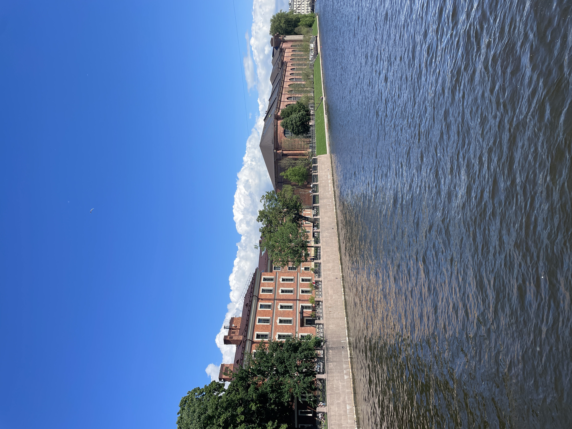 This is a medium shot of the Fontanka River in Saint Petersburg, Russia, on a clear, sunny day. On the left bank of the river, a red brick building with many windows stands adjacent to the water. Trees line the riverfront in front of the building, and a stone embankment separates the water from the sidewalk. Further down the embankment, a larger, long red brick building with a dark roof and arched windows is visible, also bordered by trees and grass. The river itself is dark blue and slightly rippled, reflecting the blue sky and some of the buildings. A few white clouds are scattered across the sky, and a single bird is visible flying in the distance.