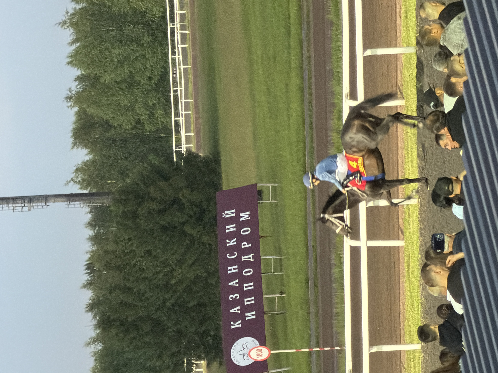 The image shows a horse race at the Kazan Hippodrome. A jockey in a blue shirt and helmet rides a dark brown horse with a red saddle pad featuring the number 4. The horse is in motion, appearing to jump or kick. In the background, a large maroon banner with white Cyrillic text reads "КАЗАНСКИЙ ИППОДРОМ". To the left of the banner, a white circular sign with a red border displays the number "1800". Spectators are visible in the foreground, seated and watching the race. The race track is dirt, with a white railing separating it from the grassy area. A tall metal structure, possibly a pylon or tower, is visible in the far background behind a line of green trees. The sky is overcast.