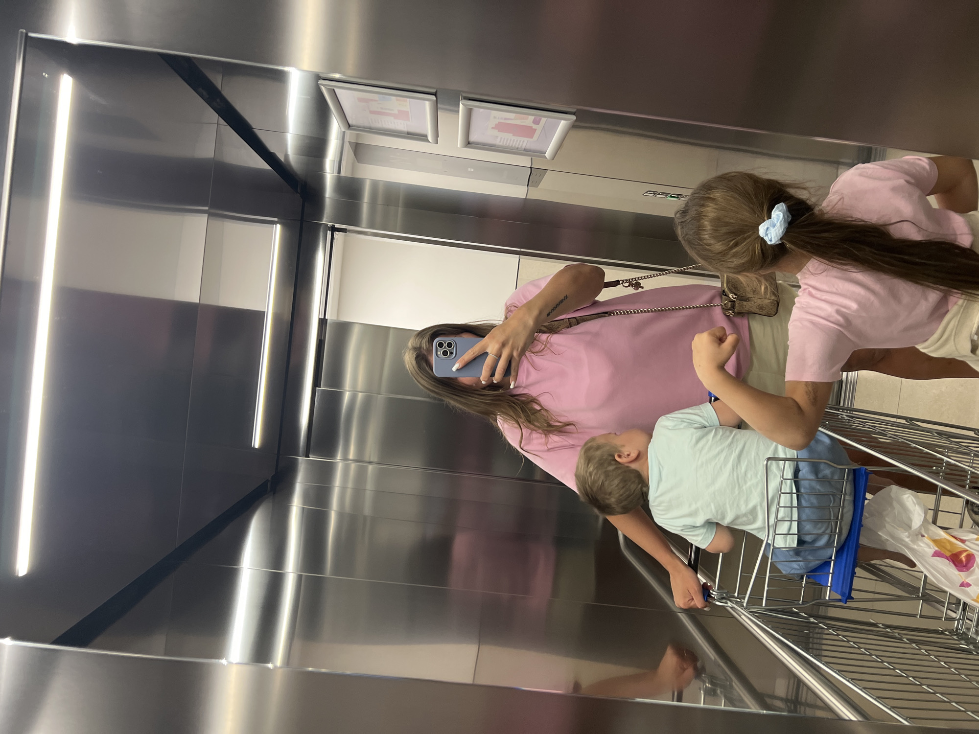 Inside an elevator with brushed metal walls, a woman in a pink shirt holds a blue smartphone, presumably taking a picture in the mirror. With her are two children: one, a girl in a pink shirt with a blue hair tie, and the other, a boy in a light blue shirt riding in a shopping cart. The location is likely a retail establishment in Budva, Montenegro.