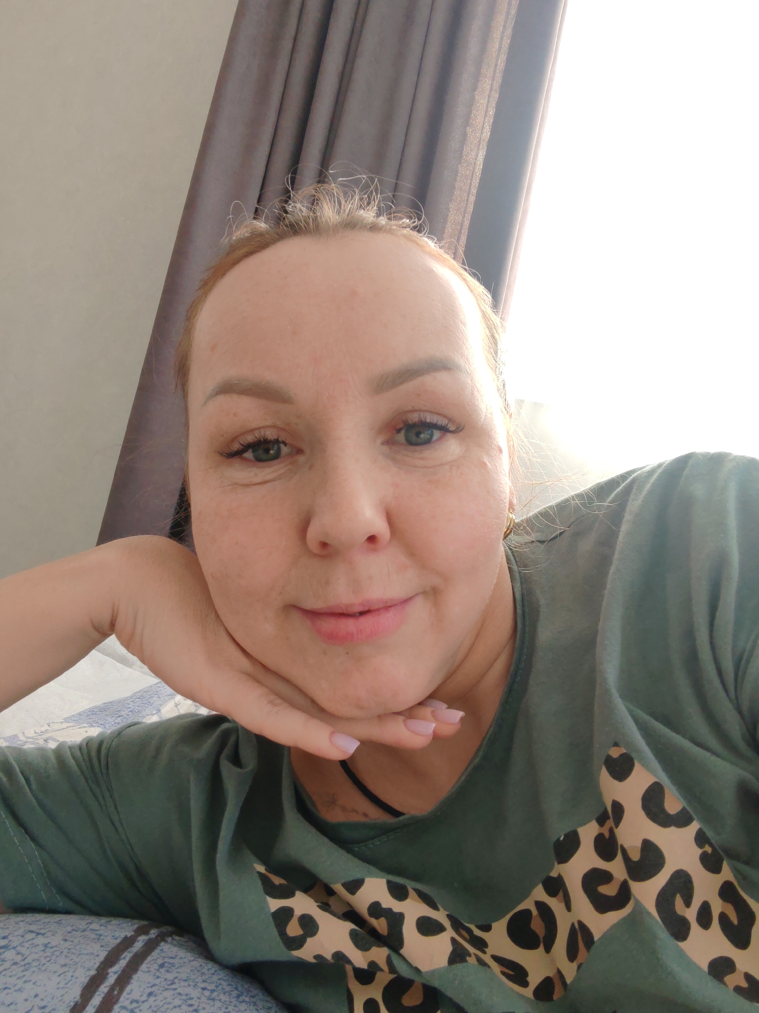 A fair-skinned adult female is depicted in a close-up, head-and-shoulders portrait, lying down and facing the camera directly. She has light reddish-brown hair, green eyes, and visible freckles on her face. Her chin rests on her right hand, which has light pink manicured fingernails. She wears a dark green t-shirt with a light brown and black leopard print design across the chest. A thin black necklace or cord is visible around her neck, and a small gold-colored hoop earring is in her right ear. The background consists of a light grey wall on the left and a drawn dark grey curtain on the right, revealing a bright, overexposed light source, consistent with a window. Blue and white patterned bedding is visible beneath her arm. The scene is located indoors, likely in a residential setting, in Chaikovsky, Russia.