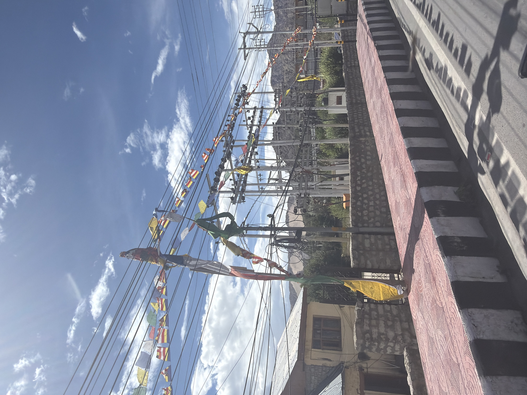 In Leh, India, prayer flags are strung across power lines above a raised sidewalk with a red tiled surface and a black and white striped border. A house with a tin roof is visible on the left, behind a stone wall. In the background, a power substation with numerous poles and wires is situated against a backdrop of barren mountains under a partly cloudy blue sky.
