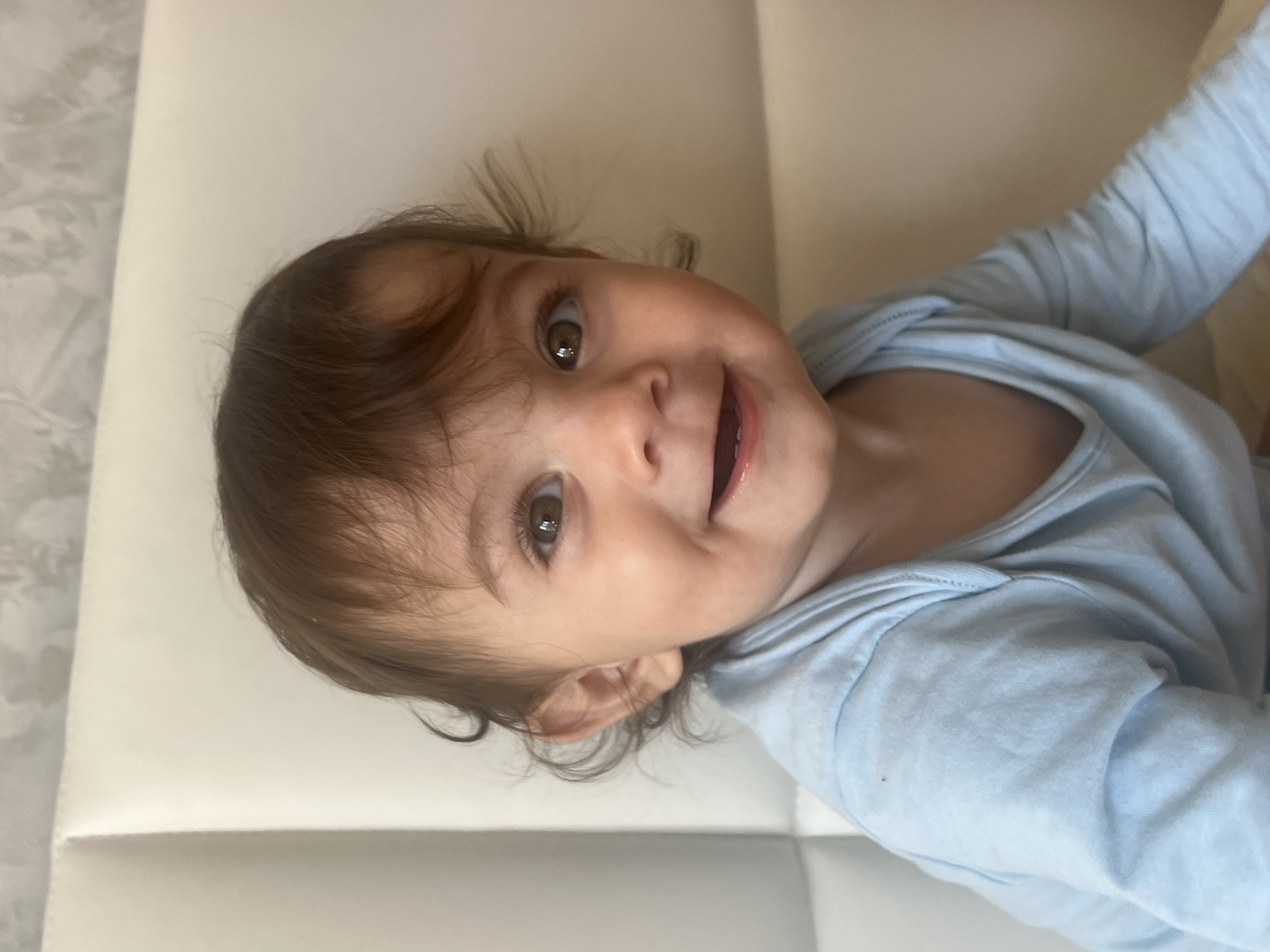 A baby with brown hair and eyes, wearing a blue long-sleeved shirt, is lying on a white surface. The scene does not provide enough information to determine the location in the city of Elena, Bulgaria.