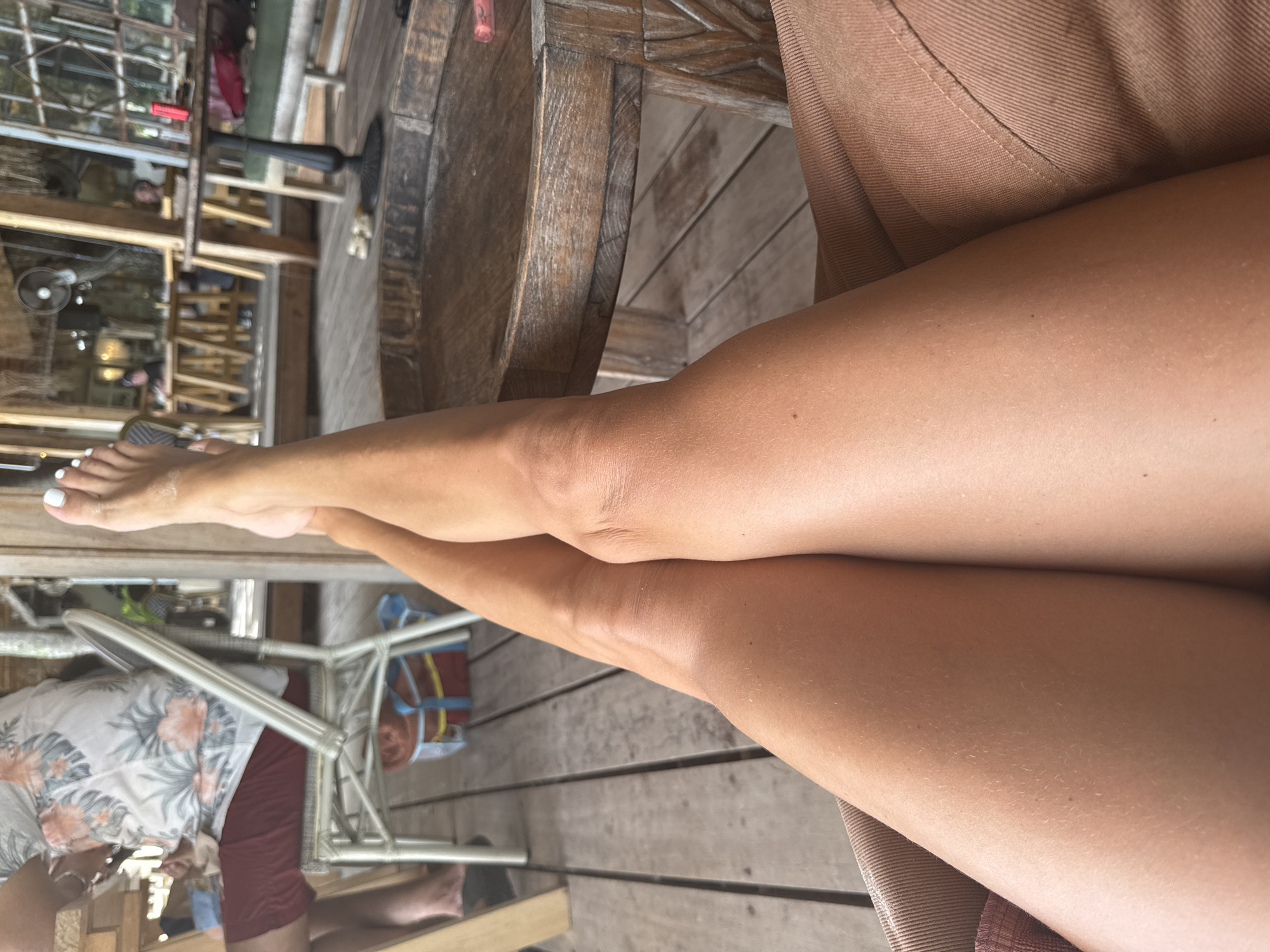 In Baan Tai, Thailand, a person's legs are positioned with their left leg crossed over their right, resting on a tan-colored cushion. Their feet, with white-painted toenails, are visible at the top of the frame. The background features a wooden deck, a circular wooden table with a carved base, and other patrons seated at tables in a dimly lit, open-air establishment. The overall ambiance suggests a casual setting, possibly a restaurant or bar.
