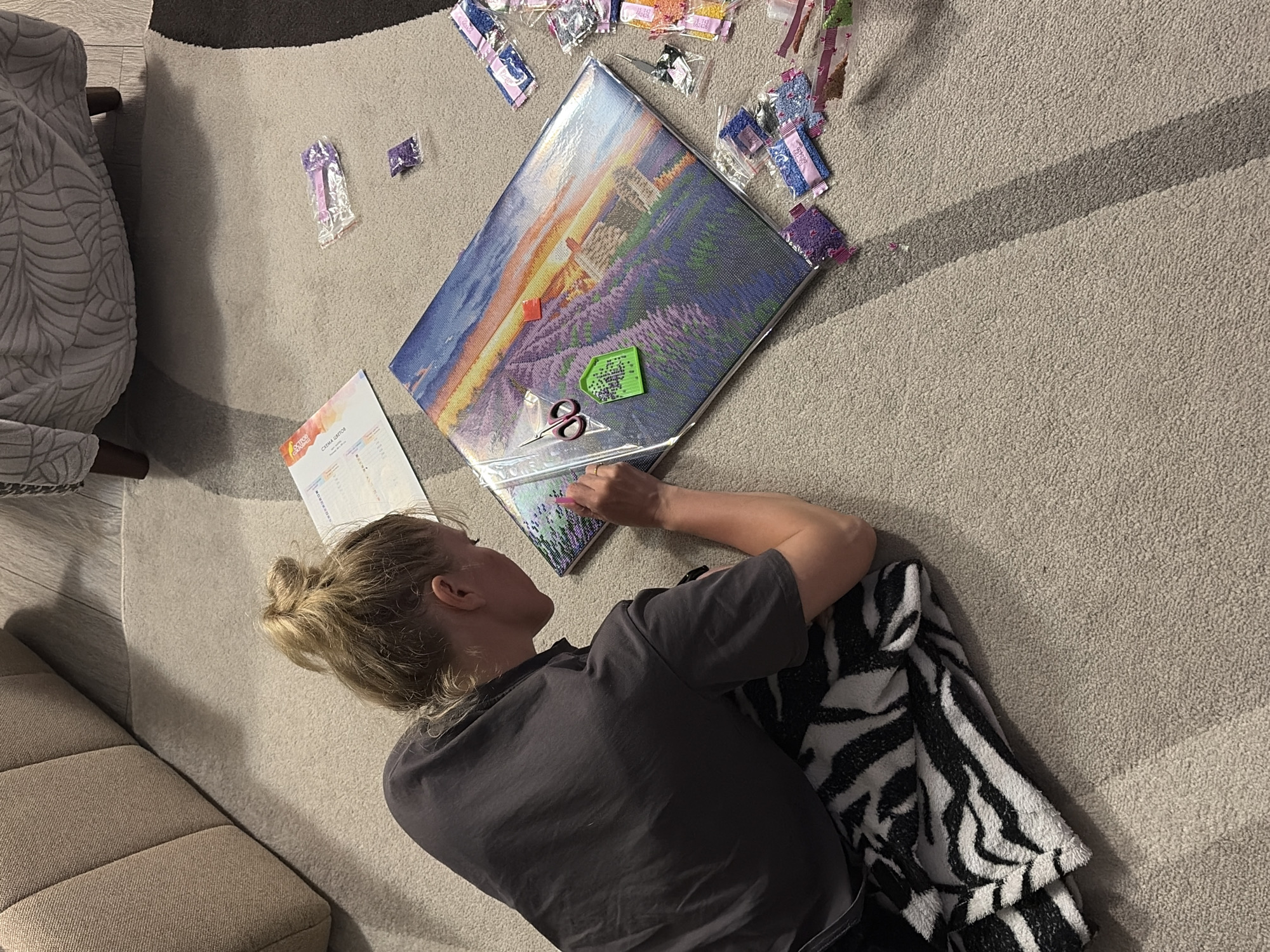 A woman with blonde hair tied in a bun is lying on a beige carpet, working on a diamond painting project. She is wearing a gray t-shirt and resting on a zebra-striped blanket. The diamond painting depicts a landscape with lavender fields, and various small plastic bags containing colored diamonds are scattered around it. A tray and a pair of scissors are placed on the painting, and a reference chart is visible to the side. The scene appears to be inside a home, and given the prompt, it is assumed to be in Chaikovsky, Russia.