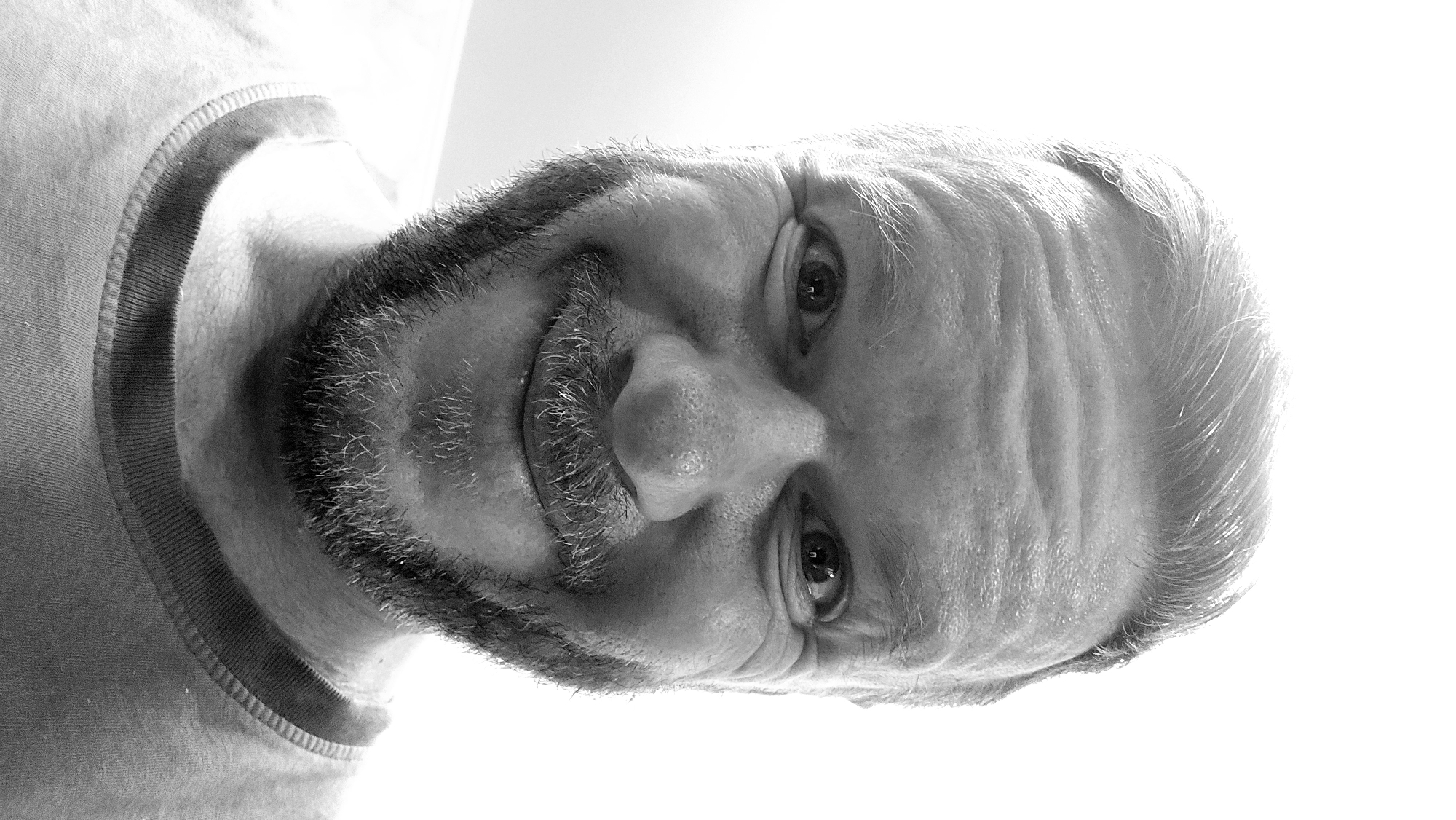 The image is a black and white, close-up portrait of a man with a short beard and mustache. He has wrinkles on his forehead and around his eyes. He is wearing a shirt with a banded neckline. The image is rotated, with the man's face tilted towards the right. There are no details in the image to indicate this photo was taken in Khimki, Russia.