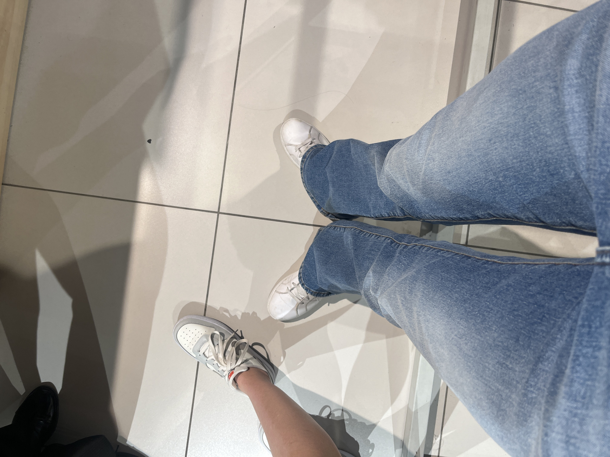 This is a first-person perspective, overhead shot of two people's legs and feet standing on a tiled floor. The person on the left has one leg visible, wearing a white and grey sneaker and blue jeans. The person on the right has both legs visible, wearing white sneakers and blue jeans. There is a shadow cast on the floor by an unseen object or person. The scene is indoors, likely a public space given the tiled floor and uniform lighting. No specific location within the city of Perm, Russia, can be identified from the image.