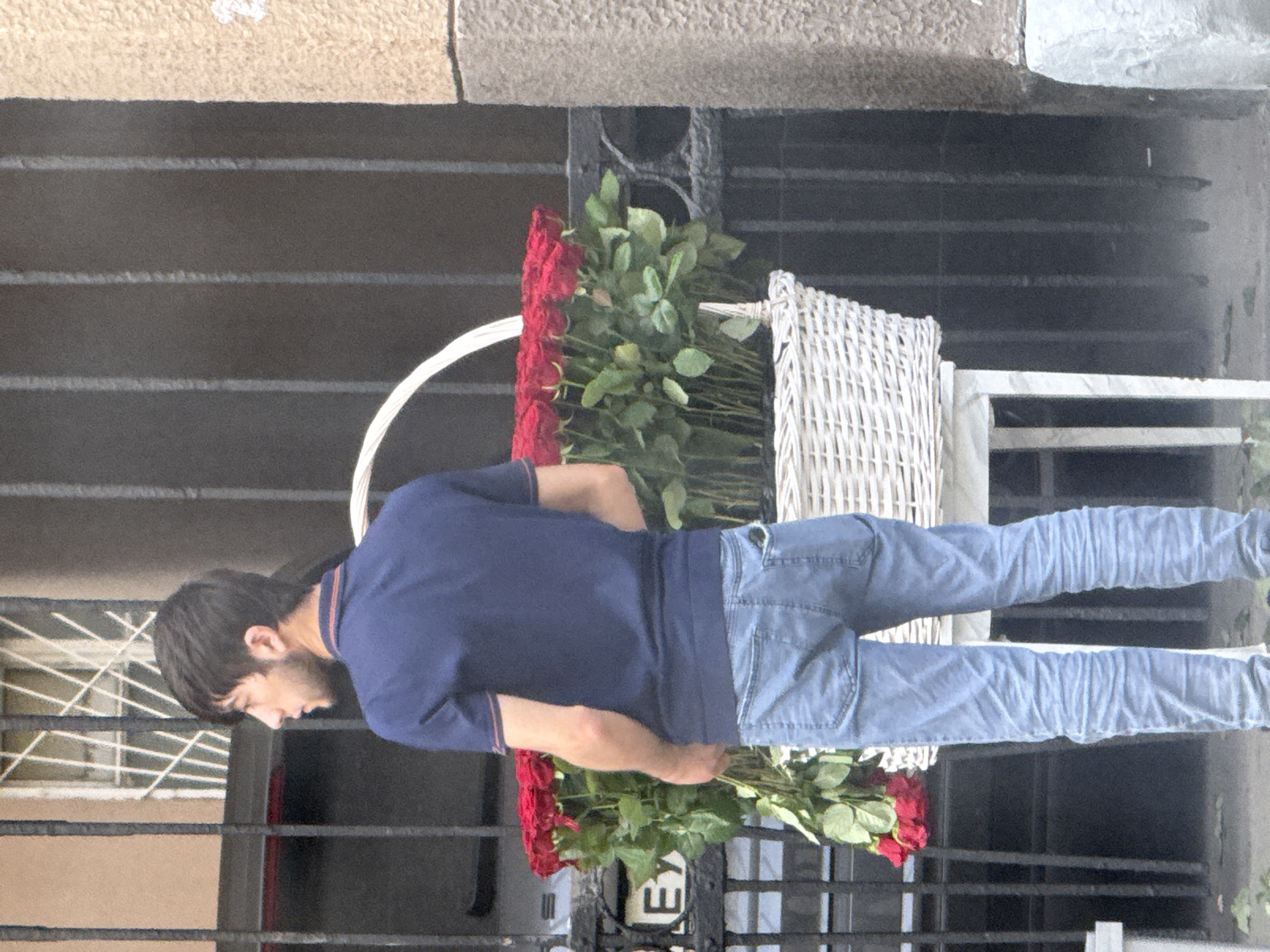 A young man stands facing away from the camera, his body angled to the left. He has dark, short hair and a small beard. He is wearing a dark blue polo shirt with red trim on the collar and sleeves, and light blue jeans. His left hand is in his pocket, and his right arm hangs loosely by his side.
In front of the man, and slightly to his right, is a large white woven basket filled with numerous red roses. The basket is elevated on a white stand. Behind the man and the roses, there is a dark, barred gate or fence. To the left of the frame, a portion of a building with a window and ornate bars is visible. The ground in front of the stand is a paved surface. The man appears to be either selling or delivering the roses. The scene suggests a street-side vendor setting or a delivery point in an urban environment. The architecture and barred gate are consistent with historical or older buildings found in cities like Moscow, Russia, where such decorative ironwork is common. The lighting suggests it is daytime.