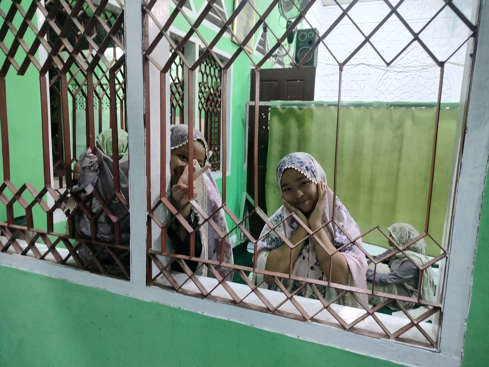 The image shows three young girls inside a building, viewed through a decorative metal grate. The building appears to be painted green on the exterior. The girls are wearing headscarves. The two girls in the foreground are smiling and looking towards the camera. One of them has her hands on her cheeks. The third girl is further back and facing away from the camera. The interior of the building is dimly lit. There is a green curtain visible behind the girls, and a speaker mounted on the wall in the background. A ceiling fan is also visible. This scene is likely a residential building or a community center in Purwodadi, Indonesia.