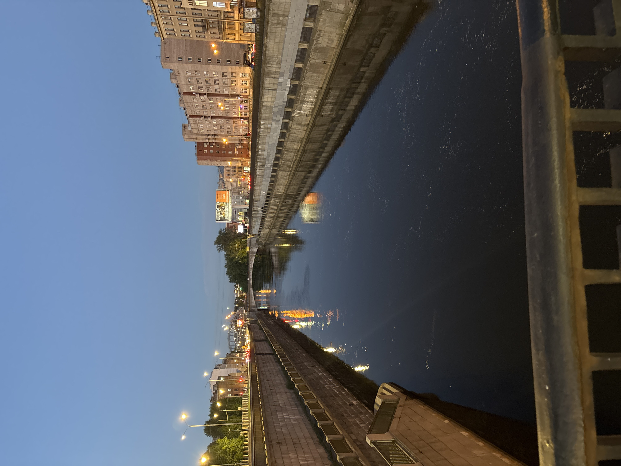 The image captures a view of the Moika River in Saint Petersburg, Russia, during dusk. The river flows through a canal lined with stone embankments. On the right bank, a series of multi-story buildings with lit windows stand in a row. On the left bank, a road with streetlights and traffic is visible, with more buildings beyond. A bridge spans the river in the distance, and lights from the city reflect on the dark water. The sky is a deep blue, indicating the transition from day to night. A metal railing is in the foreground, suggesting the photo was taken from a bridge or elevated walkway. There are no discernible people in the image.