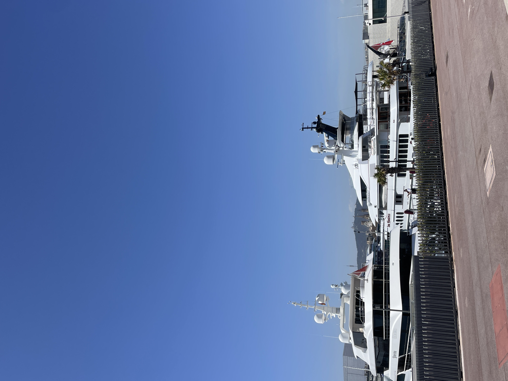 The image depicts a row of large, white yachts docked at a marina on a clear, sunny day. The sky is a deep blue, taking up a large portion of the frame. The yachts are lined up along a quay, separated from the walkway by a dark gray fence. A red sidewalk runs alongside the yachts and the fence. The names "Sea Huntress" and "Severka" are visible on two of the boats. A few palm trees are seen between the boats. The backdrop is blurred, but appears to be a cityscape, hinting at a coastal urban area. The location is potentially Barcelona, Spain.