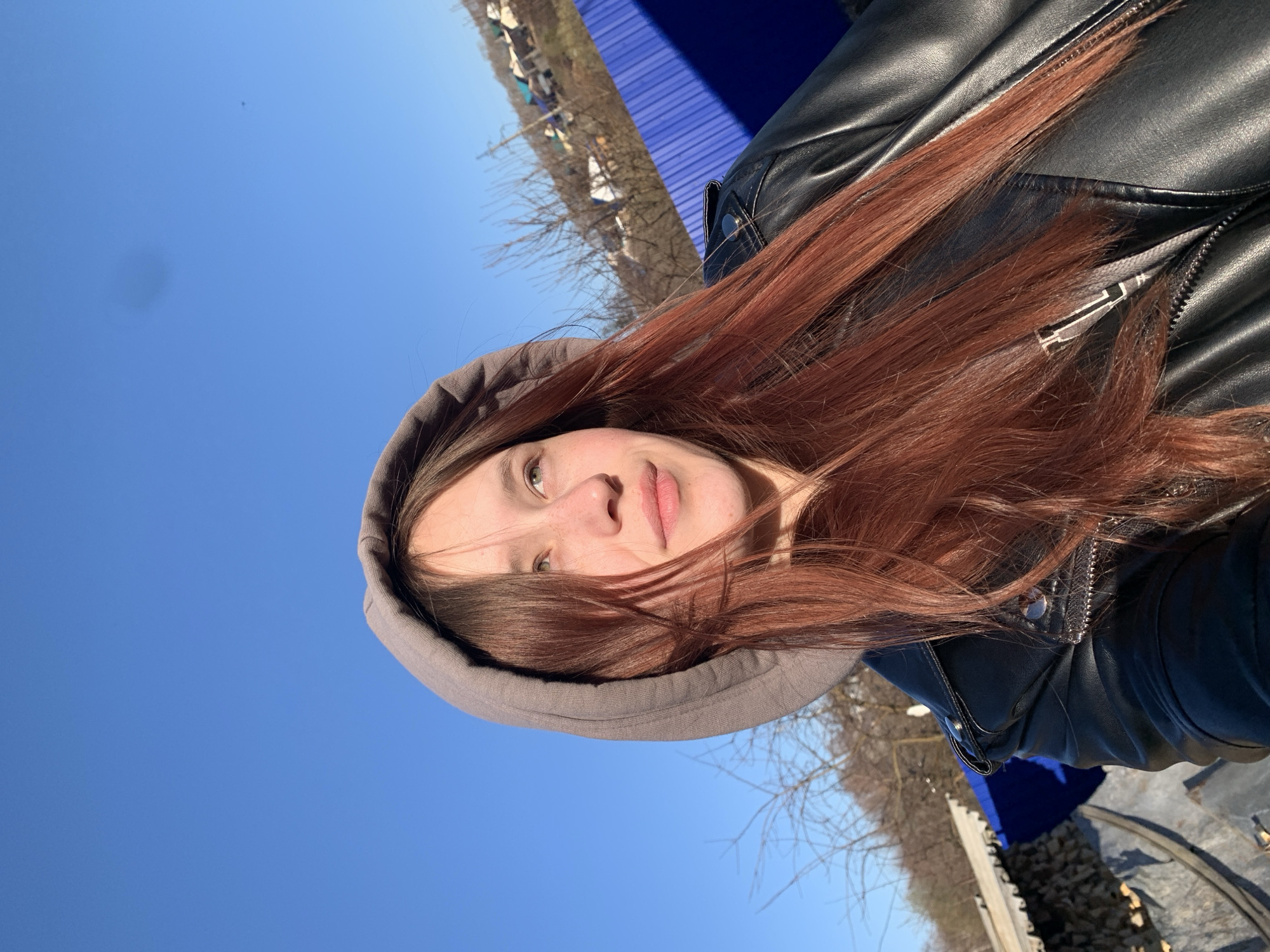 A young woman is captured in a sunlit selfie, looking directly at the viewer with a calm, neutral expression and a faint smile. She has long, reddish-brown hair, bright green eyes, and visible freckles on her nose and cheeks. She is wearing a grey hooded top, with the hood pulled up, underneath a black leather jacket featuring zippers and snap buttons. A white tag with illegible black text is partially visible on the inside of her jacket.
The setting is outdoors on a bright, clear day, likely in Фоки, Russia. A vast, deep blue sky fills the upper portion of the background, indicating sunny weather. In the mid-ground and to the right, bare trees, characteristic of late winter or early spring, are visible against the sky, alongside some sparse green foliage. A prominent dark blue corrugated structure or fence appears to the right of the woman. Further in the distance, some indistinct structures, possibly tents, can be faintly seen. The bottom right corner shows a pile of wood chips or gravel and a partial circular metal object. The strong, direct sunlight illuminates the woman's face.