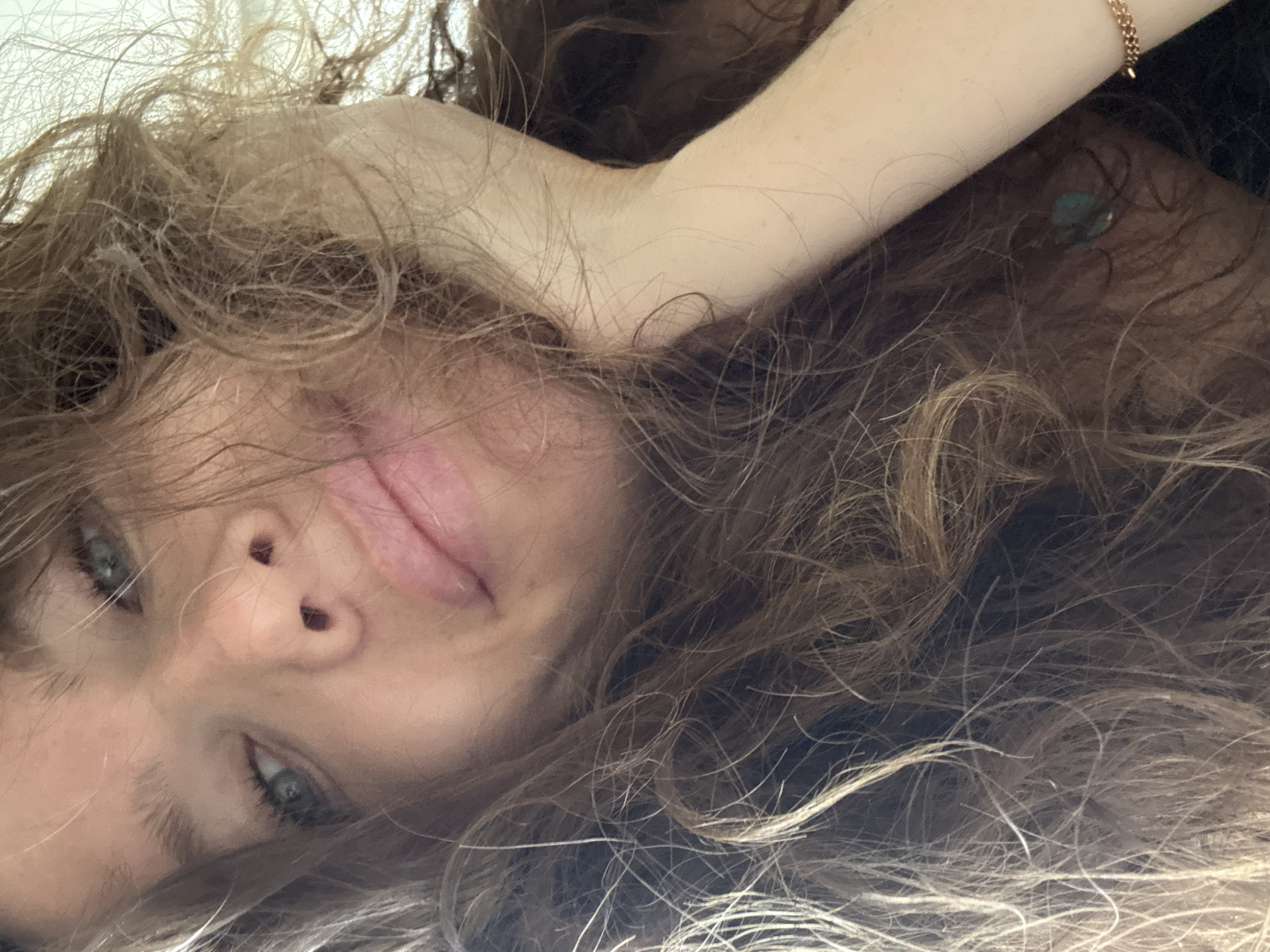 A close-up, rotated portrait shows a fair-skinned person with light eyes and light brown, wavy hair. The person is lying down. An arm is visible above the person's head, wearing a thin gold bracelet. A turquoise or blue earring is visible behind the arm. The background is dark and indistinct. There is no information related to the city of Khimki, Russia, in this image.