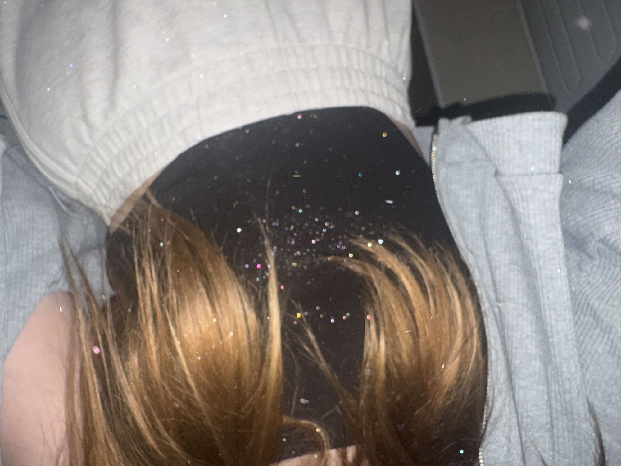This is a close-up, waist-up indoor shot of a person wearing a black crop top and light-colored sweatpants, with a grey hooded sweatshirt draped over their shoulders. The person has long, light brown hair with streaks of lighter blonde. The clothing and hair are dusted with fine, multi-colored glitter. The background is dark and out of focus. There is no indication of the city of Chaikovsky, Russia, or any other specific location. The image appears to be a self-portrait taken in an indoor setting.