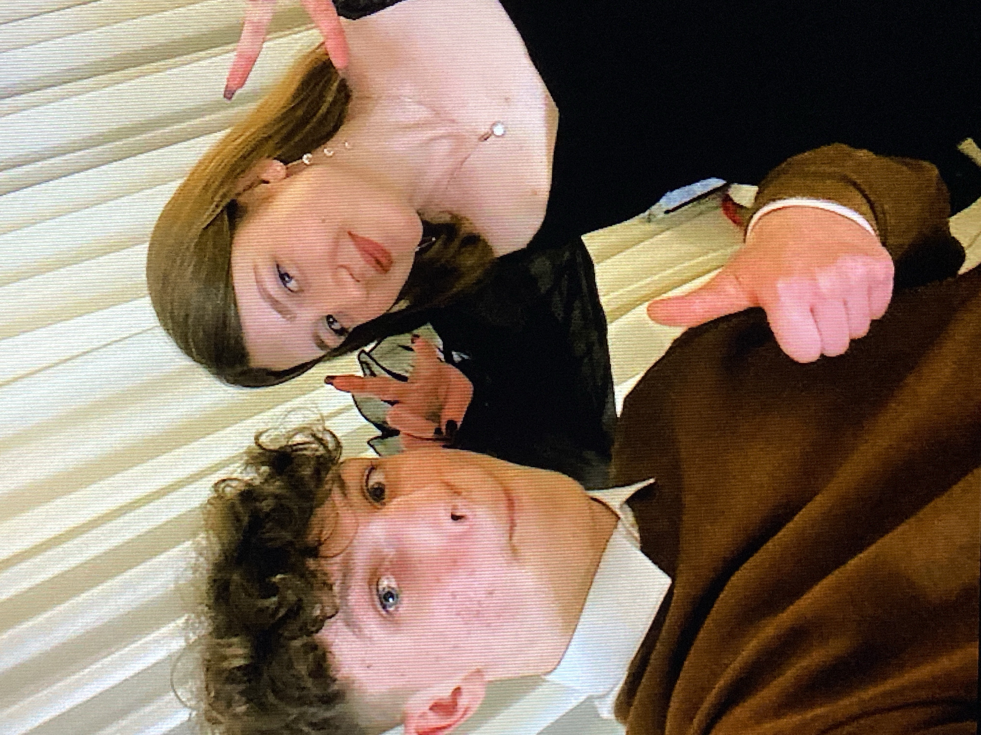 Two young adults, a male and a female, pose for a selfie in front of a cream-colored, pleated curtain, possibly in a room within the city of Poznan, Poland. The male, on the left, has short, curly brown hair and is wearing a brown sweater over a white collared shirt. He is looking directly at the camera with a slight smile and holding up a thumb in a "thumbs up" gesture. The female, on the right, has long brown hair and is wearing a black dress. She is looking towards the camera and holding up her hand in a peace sign. Both individuals appear to be in their late teens or early twenties. The lighting in the image is somewhat dim, suggesting an indoor setting.