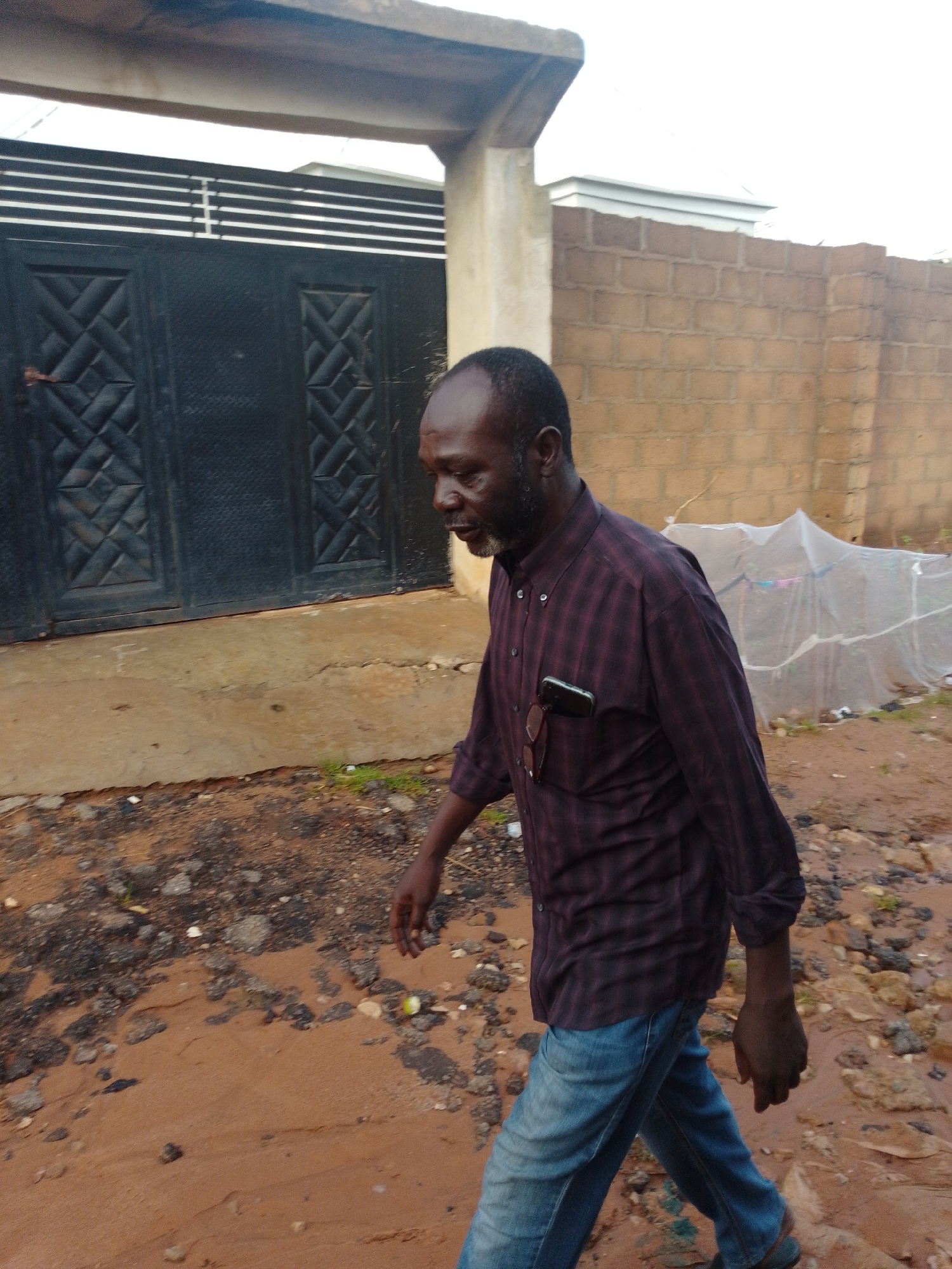 A man with dark skin, a beard, and a bald head walks to the right in front of a large black gate. He is wearing a long-sleeved, dark red plaid shirt tucked into blue jeans. Sunglasses are tucked into his shirt pocket, and a black smartphone is also in his pocket. The man is walking on a dirt path with scattered rocks and debris. Behind him, a brick wall borders the property, and a structure covered in white plastic sheeting is visible. The area appears to be an outdoor, possibly semi-urban or rural, environment.