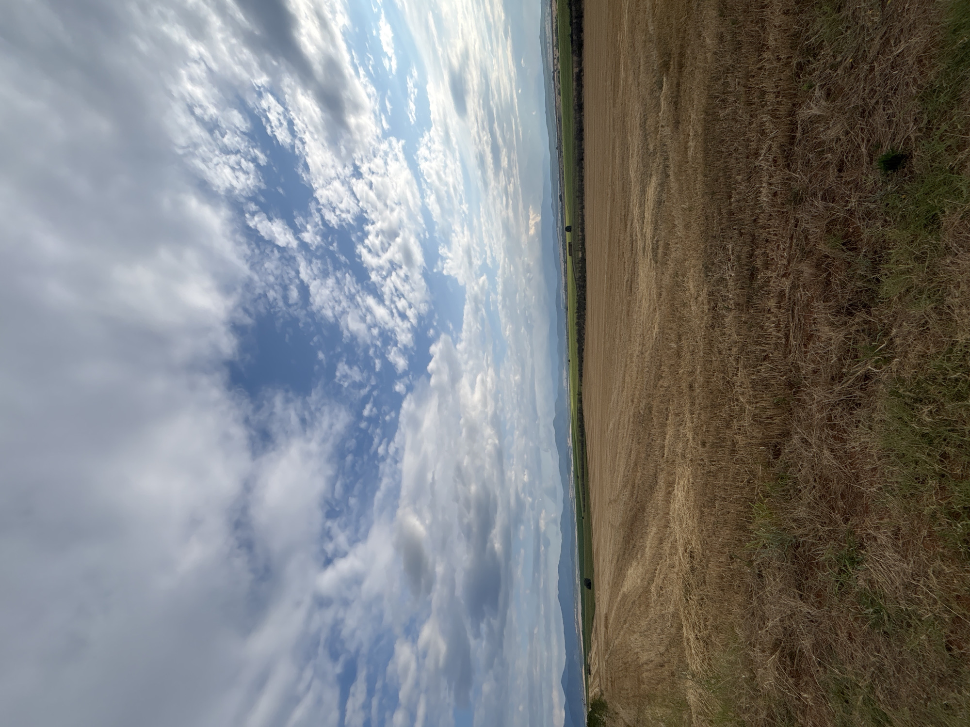 The image depicts a rural landscape under a cloudy sky, characteristic of the region surrounding Hisarya, Bulgaria. In the foreground, a field of harvested golden wheat stubble stretches horizontally across the frame. The stubble shows the patterns of agricultural machinery, indicating recent harvesting. The middle ground features a transition to greener fields, with distant trees and what appear to be hay bales or agricultural structures. Further in the distance, a range of mountains forms the horizon, silhouetted against the sky. The sky itself is dynamic, with a mix of white and grey clouds interspersed with patches of blue. The overall scene is one of agricultural land, likely during late summer or early autumn, with a natural, serene ambiance. There are no people or animals visible in the image.