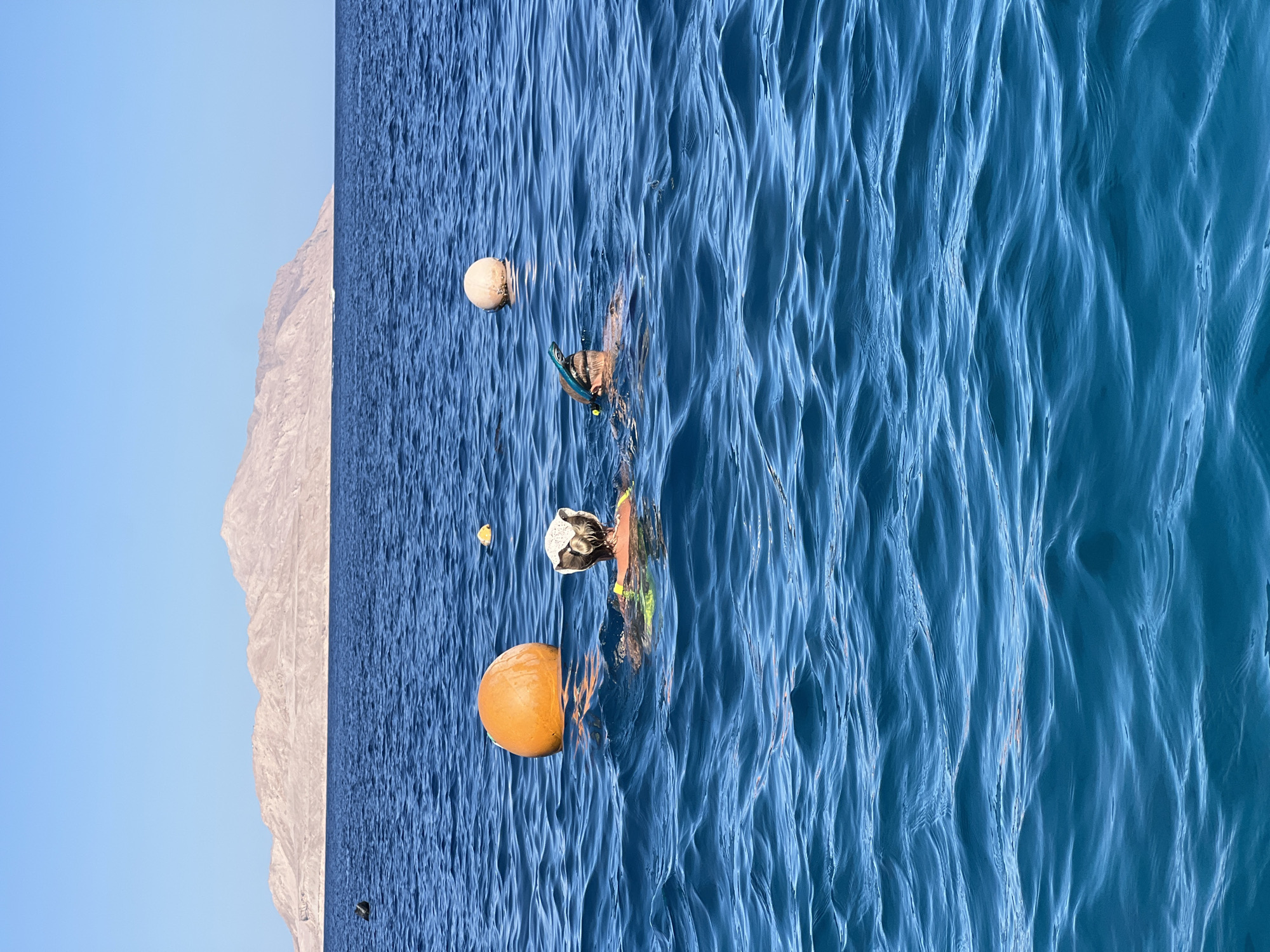 Two individuals are snorkeling in the clear blue waters of Sharm El Sheikh, Egypt, with a rugged, mountainous island in the background under a clear sky. An orange buoy is positioned to the left of the two snorkelers, and a smaller white buoy is further to the right. One snorkeler, wearing a yellow vest, faces away from the camera, with a white cap on their head. The other snorkeler, partially obscured, wears a snorkel mask and is looking towards the island. The ocean surface is rippled with gentle waves.