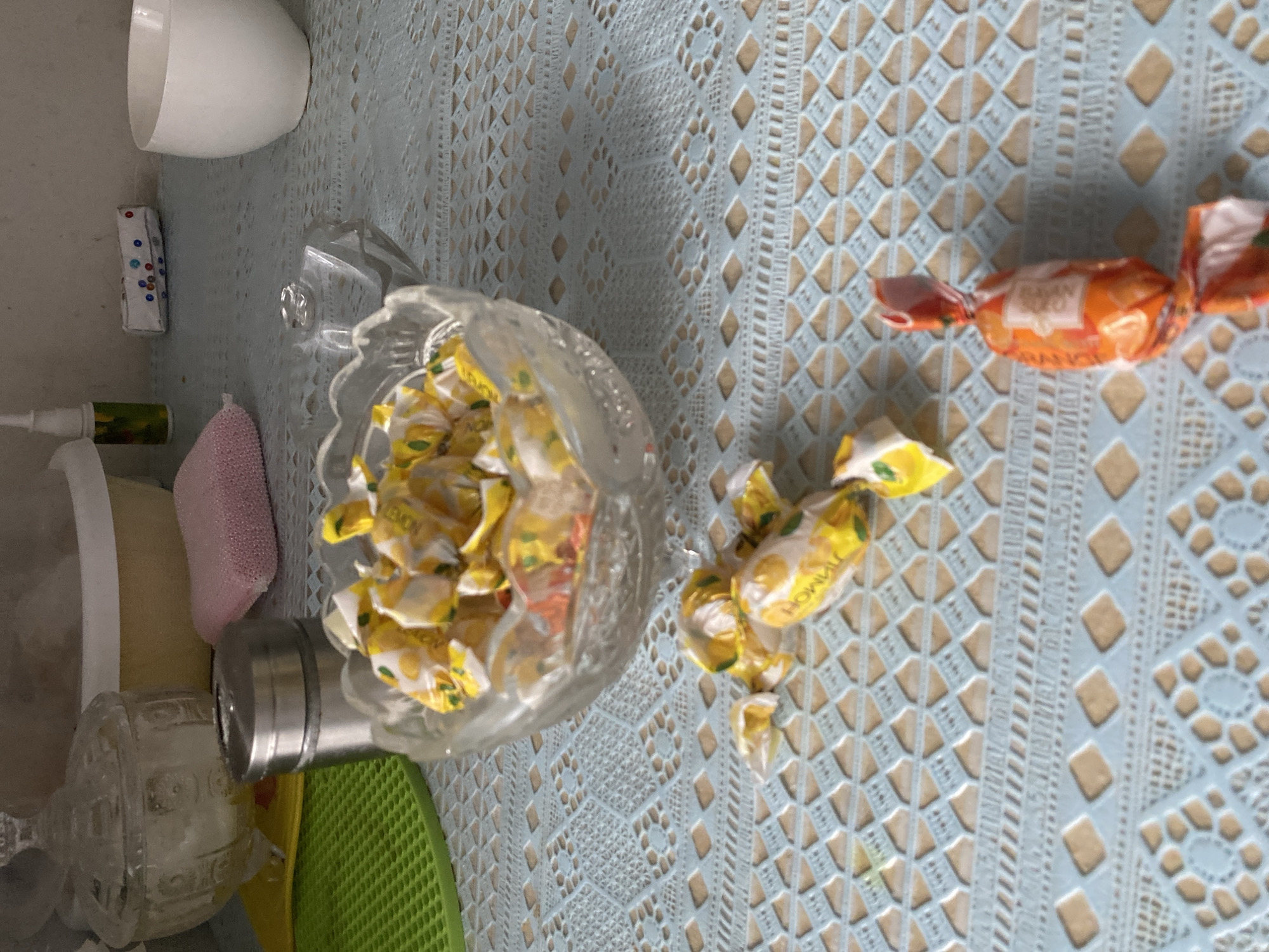 The image depicts a close-up, overhead view of a table with a patterned light blue tablecloth. Several candies are scattered across the tablecloth. In the center, a glass bowl is filled with wrapped yellow and white candies, some of which appear to be lemon-flavored. To the right of the bowl, two individually wrapped candies lie on the tablecloth: one is orange and white, and the other is orange with some white elements. To the left of the bowl, there are more scattered yellow and white wrapped candies. In the background, to the left, a clear glass sugar bowl with a lid is visible, next to a stainless steel container, and a pink sponge. Further back, a white ceramic bowl and a small bottle with a dispenser are present. The setting appears to be a domestic indoor environment. The specific location in Petropavl, Kazakhstan, cannot be determined from the image alone.
