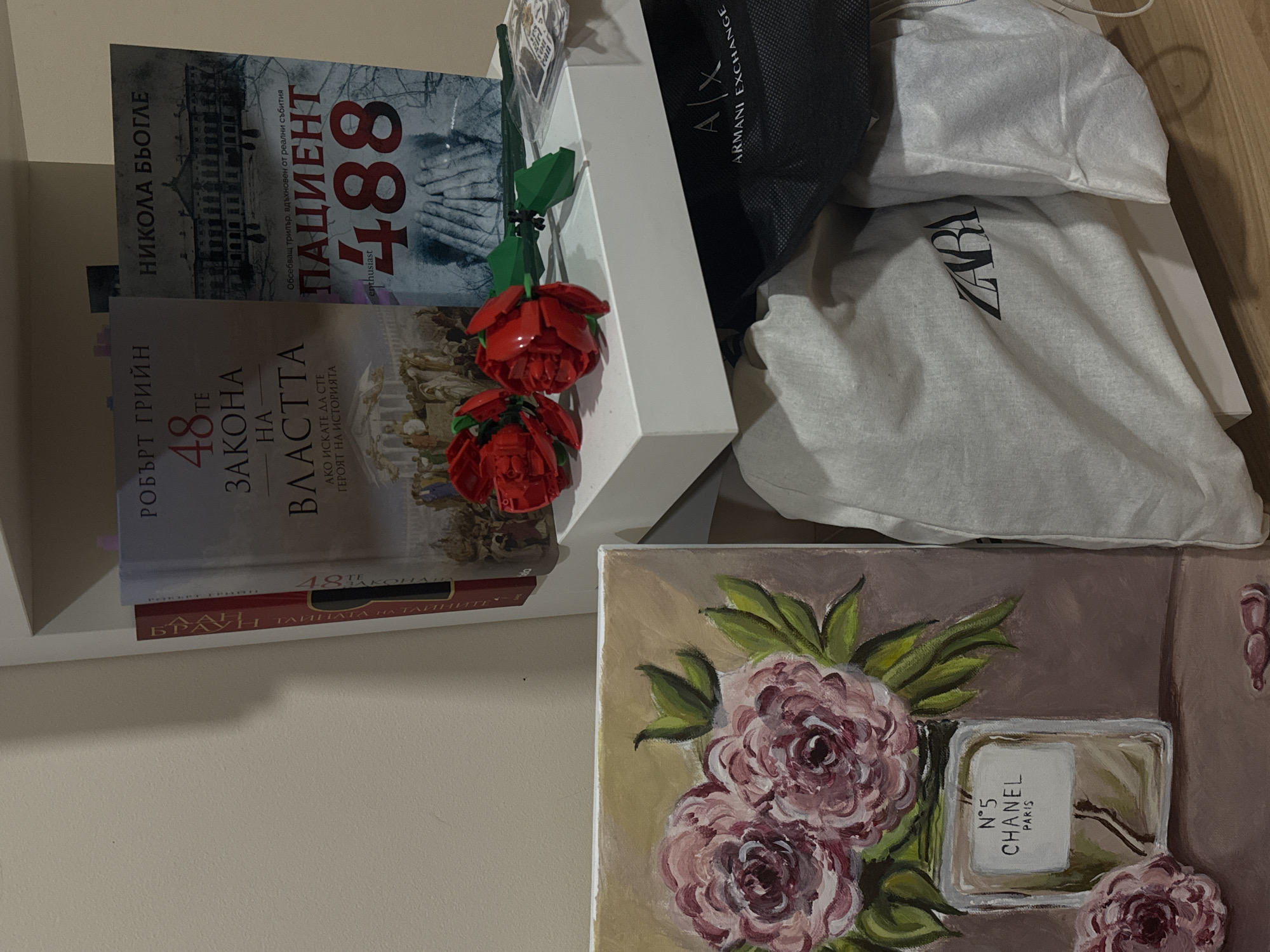 The image displays an indoor scene, likely in a residential setting in Sofia, Bulgaria, as indicated by the Bulgarian text on the books. A white shelving unit occupies the right side of the frame, with items placed on its top surface and below.
On the upper surface of the shelf, three books are arranged vertically. From left to right: a book with a red spine partially visible, identifiable as "БРАУН ТАЙНАТА НА ДА ВИНЧИ" (likely Dan Brown's "The Da Vinci Code"); "РОБЪРТ ГРИЙН 48-ТЕ ЗАКОНА НА ВЛАСТТА" (Robert Greene's "The 48 Laws of Power"); and "НИКОЛА БЬОГЛЕ ПАЦИЕНТ 488" (Nicolas Beuglet's "Patient 488"). In front of these books, two red, plastic, brick-built roses with green stems lie horizontally. A small white sticker with the phrase "you go girl you can do it" in black text is also visible near the roses.
Below the shelf, a black mesh bag featuring the "A|X ARMANI EXCHANGE" logo in white is present, partially obscured. Beside it are a crumpled light grey fabric bag with the "ZARA" logo in black, and another crumpled white fabric bag. These items suggest storage or recent acquisition.
To the left of the shelf unit, a rectangular canvas painting is propped vertically. The painting depicts three pinkish-purple roses with green leaves, arranged above a stylized rectangular bottle labeled "N°5 CHANEL PARIS" in white text. The background of the painting is a solid light brown hue. On the surface directly below the painting, a small, glossy, pink, decorative object, possibly resembling a link, is visible.
The overall environment features a plain light beige wall behind the displayed items and a light-colored wooden or laminate floor in the foreground.
