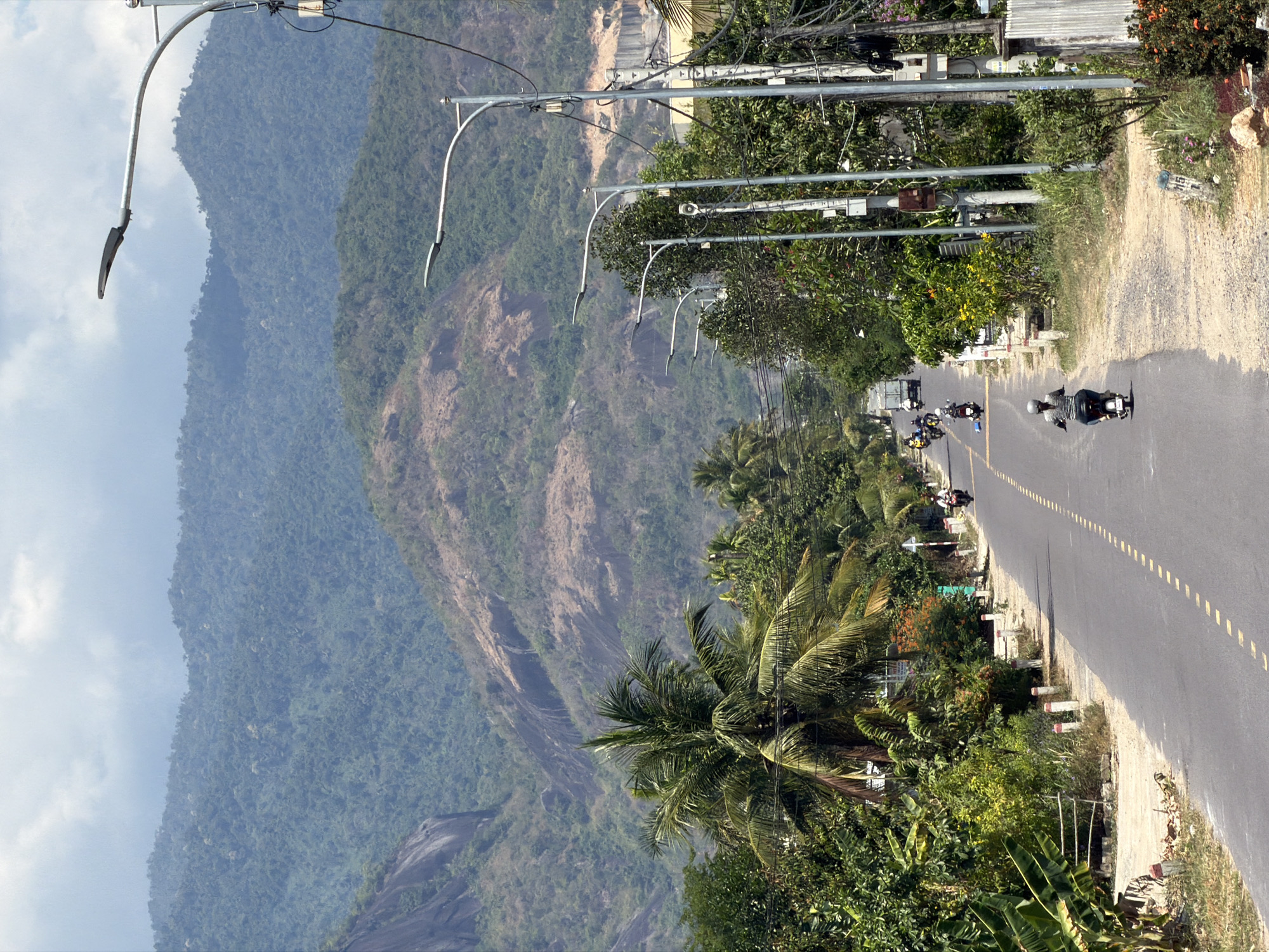 The image shows a view of a road in Diên Thọ, Vietnam, with mountains in the background. The road has a dashed yellow line down the middle and motorcycles are driving on it. There are palm trees and other vegetation along the roadside. Streetlights are visible along the road. The mountain range on the left is densely vegetated. The road is paved, and there's a dirt shoulder. Some small buildings or structures are visible among the trees along the road.