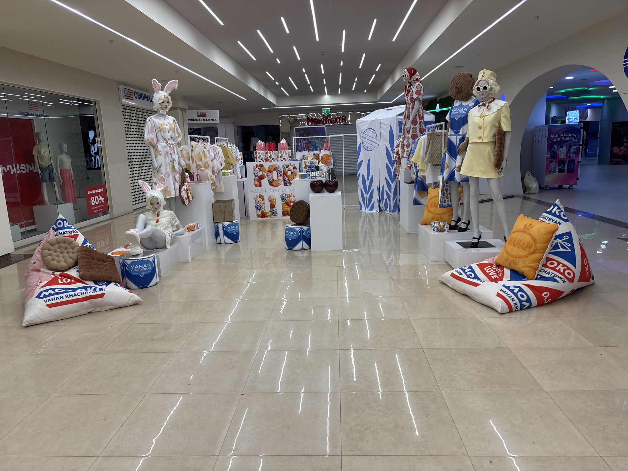 The image shows a display inside a shopping mall in Yerevan, Armenia. It is an Easter-themed display featuring mannequins dressed in clothing, including one dressed as a bunny. The mannequins are arranged on white platforms and surrounded by branded pillows. Storefronts are visible in the background.