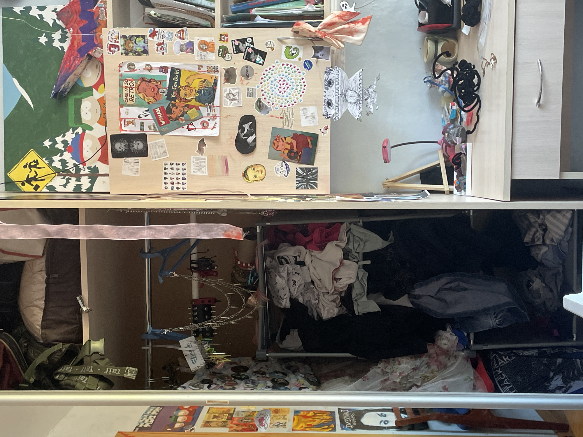 The image shows a chaotic storage space. The top shelf is cluttered with various items, including artwork featuring characters resembling those from the "South Park" animated series, and a board covered in stickers and ephemera. Other items include headwear, and various odds and ends.
Below, the lower shelf is crammed with clothing and accessories, including what appears to be a bag, hanging garments, and assorted personal items. A drawer partially obscures the view below, but what is visible reveals a chaotic arrangement of art and personal objects. A small tag is visible that says "Taif" which is the name of the city of Siva, Russia, therefore this is most likely the location of the storage space.