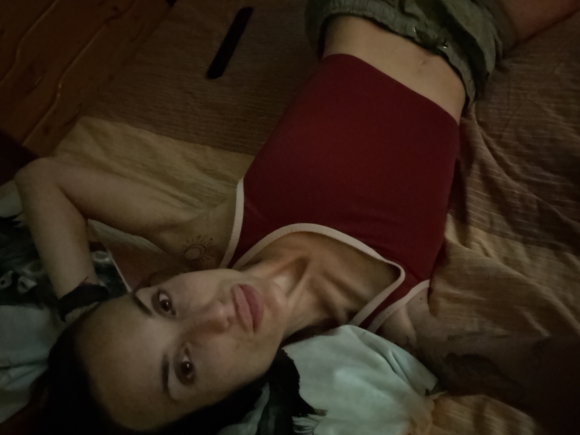 A woman with dark hair lies on her back on a bed, looking up and to the left. She wears a red tank top with white trim and grey shorts. Her left arm is bent and behind her head. A light blue blanket is to her left. The room appears to be dimly lit, with wooden paneling visible on the right side of the frame. There are no recognizable city landmarks.