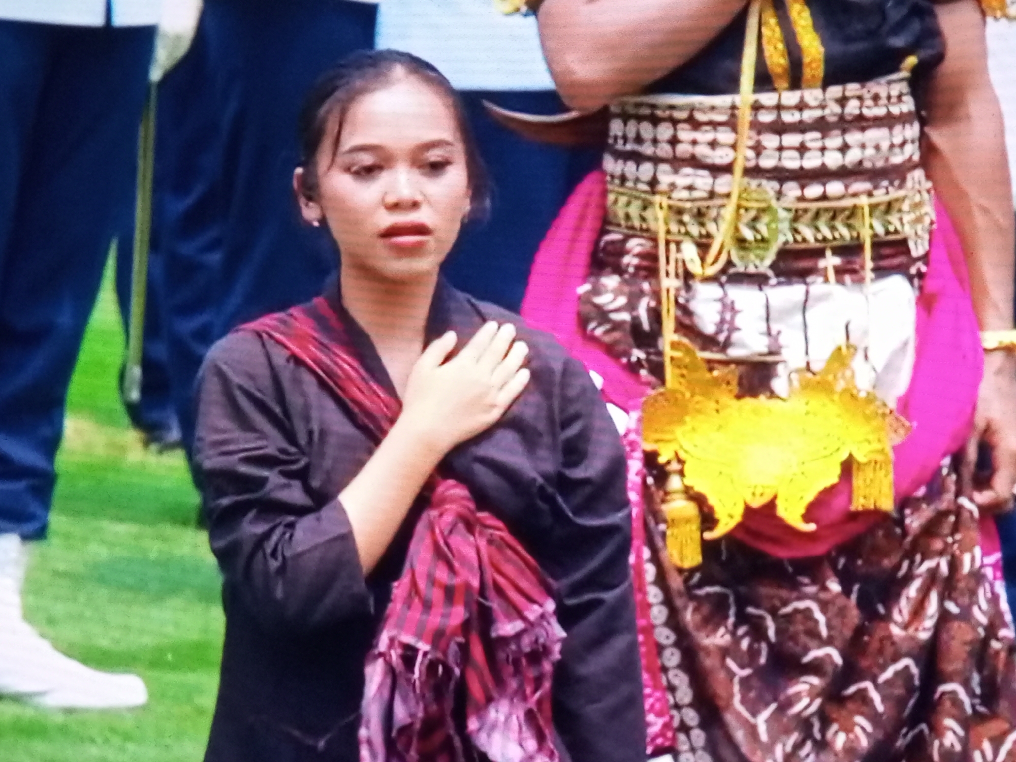 In Banyudono, Indonesia, a young woman stands on a grassy area. She is wearing a dark top with a red and black striped scarf draped around her neck and shoulders, with one end of the scarf resting on her chest. Her right hand is placed over her heart. Behind her, a person is wearing a traditional Indonesian costume, featuring a gold and beaded belt, a pink sash, and a patterned skirt. The background consists of blurred figures and dark blue fabric.