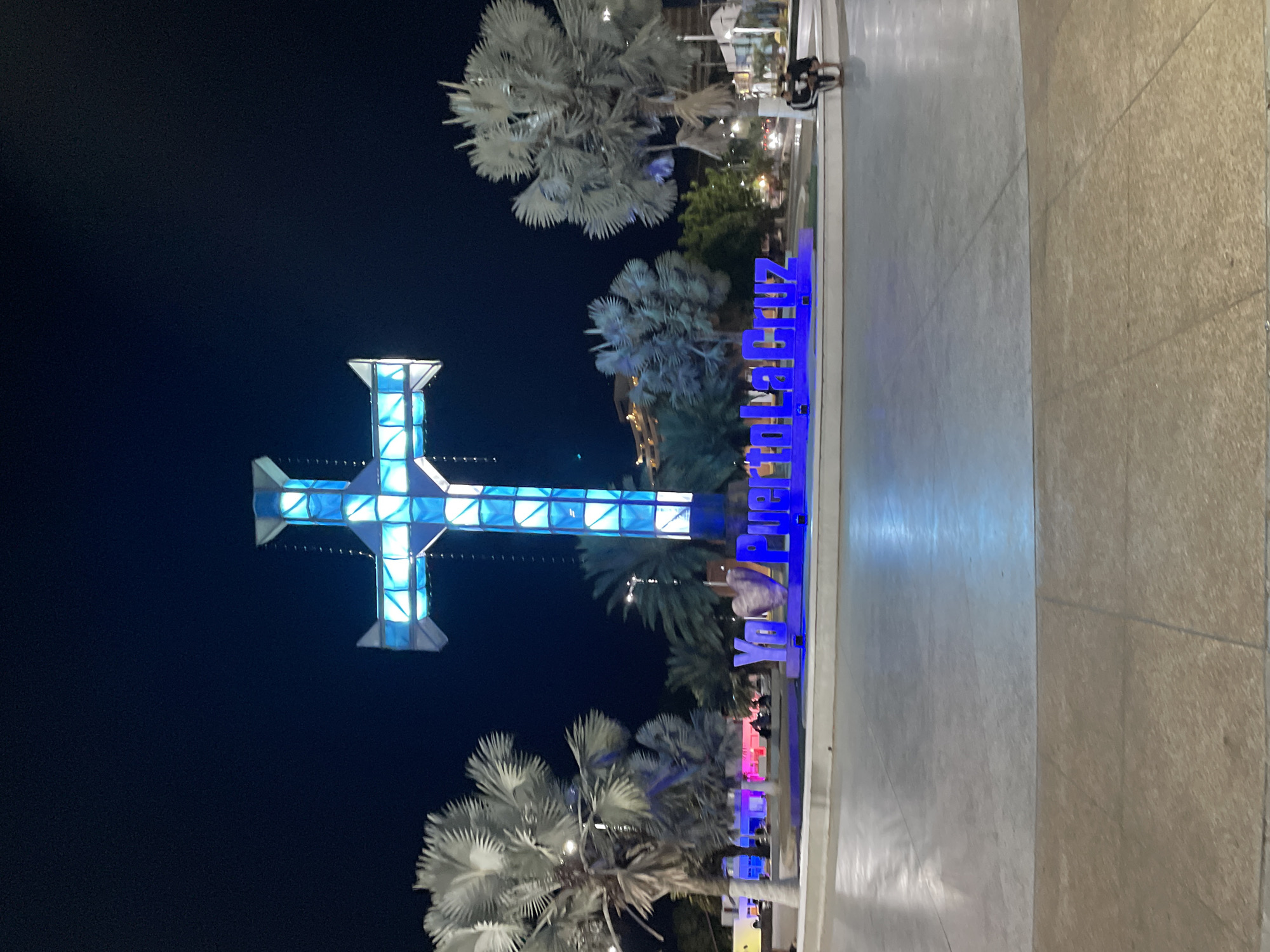 The image captures an outdoor public space at night, dominated by two brightly illuminated structures. On the left, a large, prominent cross is visible, constructed from clear or translucent panels and glowing with vibrant blue and white light. To the right of the cross, a long, illuminated sign spells out "PUERTO LA CRUZ" in large, blue capital letters, confirming the location as Puerto La Cruz, Venezuela.
Several tropical palm trees with large, light-colored fronds are scattered throughout the scene, some appearing near the cross and others around the "PUERTO LA CRUZ" sign. The ground in the foreground is a wide, light-colored paved or tiled area, resembling a plaza or promenade. In the background, a few indistinct figures can be seen walking or standing, indicating human activity in the area. The sky is dark, characteristic of nighttime, with the artificial lights of the cross and the sign standing out prominently. The scene suggests a well-lit and active public square or landmark area during the evening.