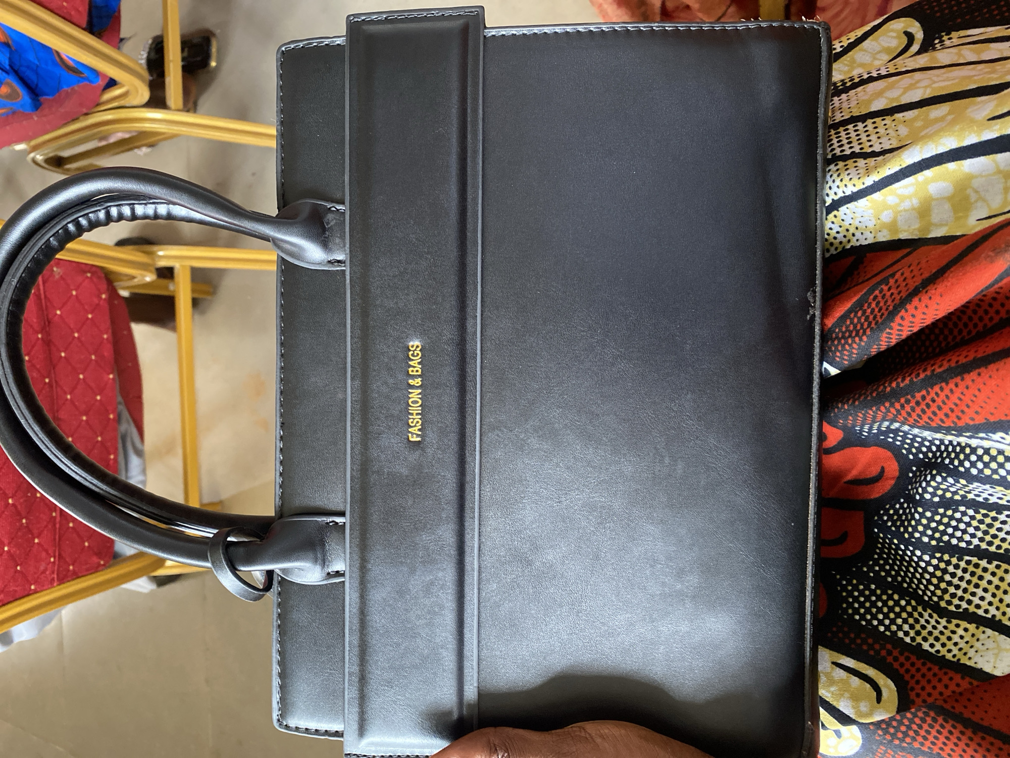 A black handbag with gold lettering that reads "FASHION & BAGS" is held by a person's left hand. The handbag is resting on a colorful fabric. In the background, red chairs with yellow frames are visible, suggesting an indoor setting. The scene is likely in Iddah, Nigeria.