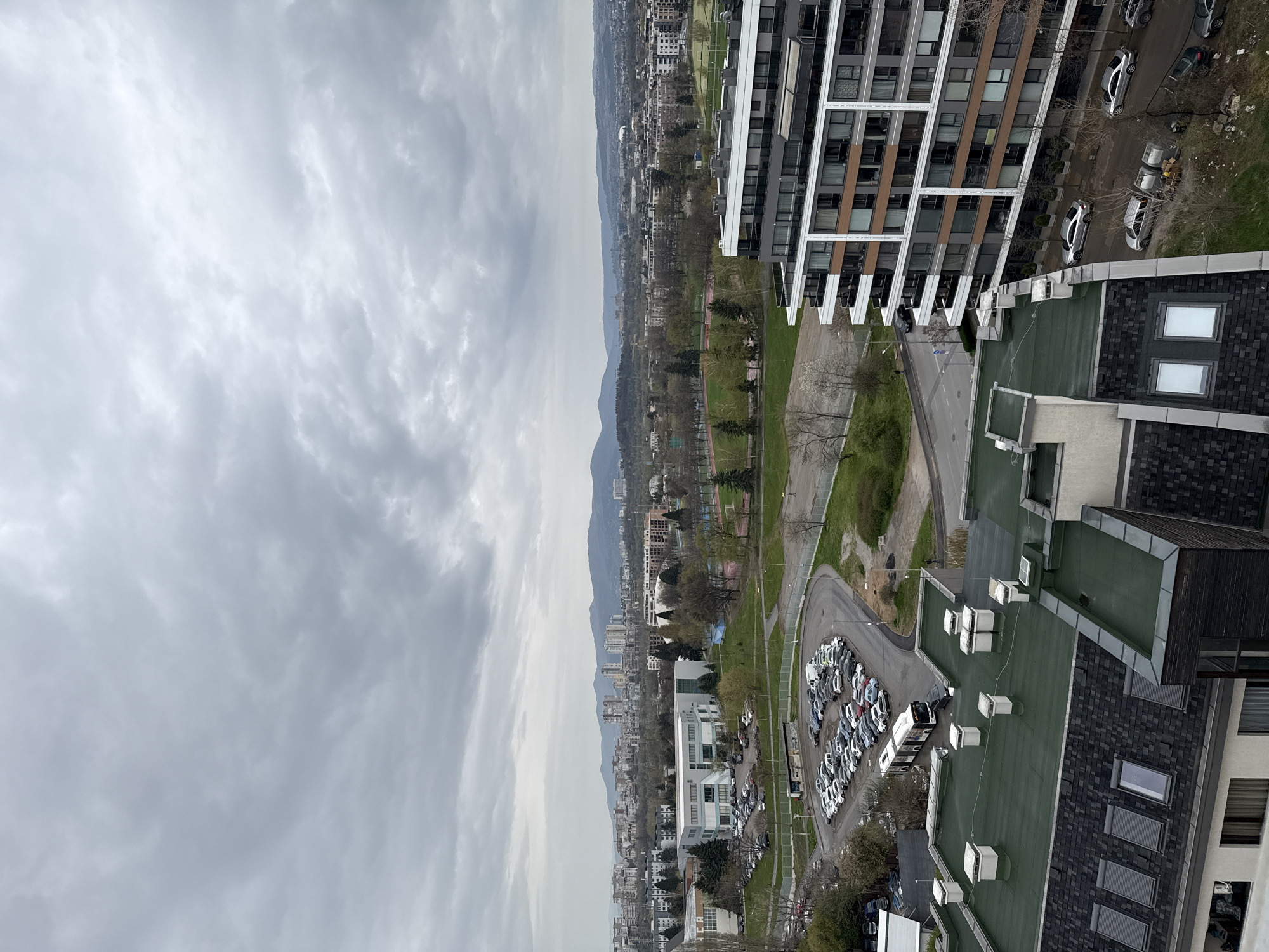 An aerial shot captures a sprawling cityscape under a cloudy sky. The built environment is a mix of residential and commercial structures, some with visible parking areas containing multiple vehicles. The location is Sofia, Bulgaria. Mountains are seen in the distance.