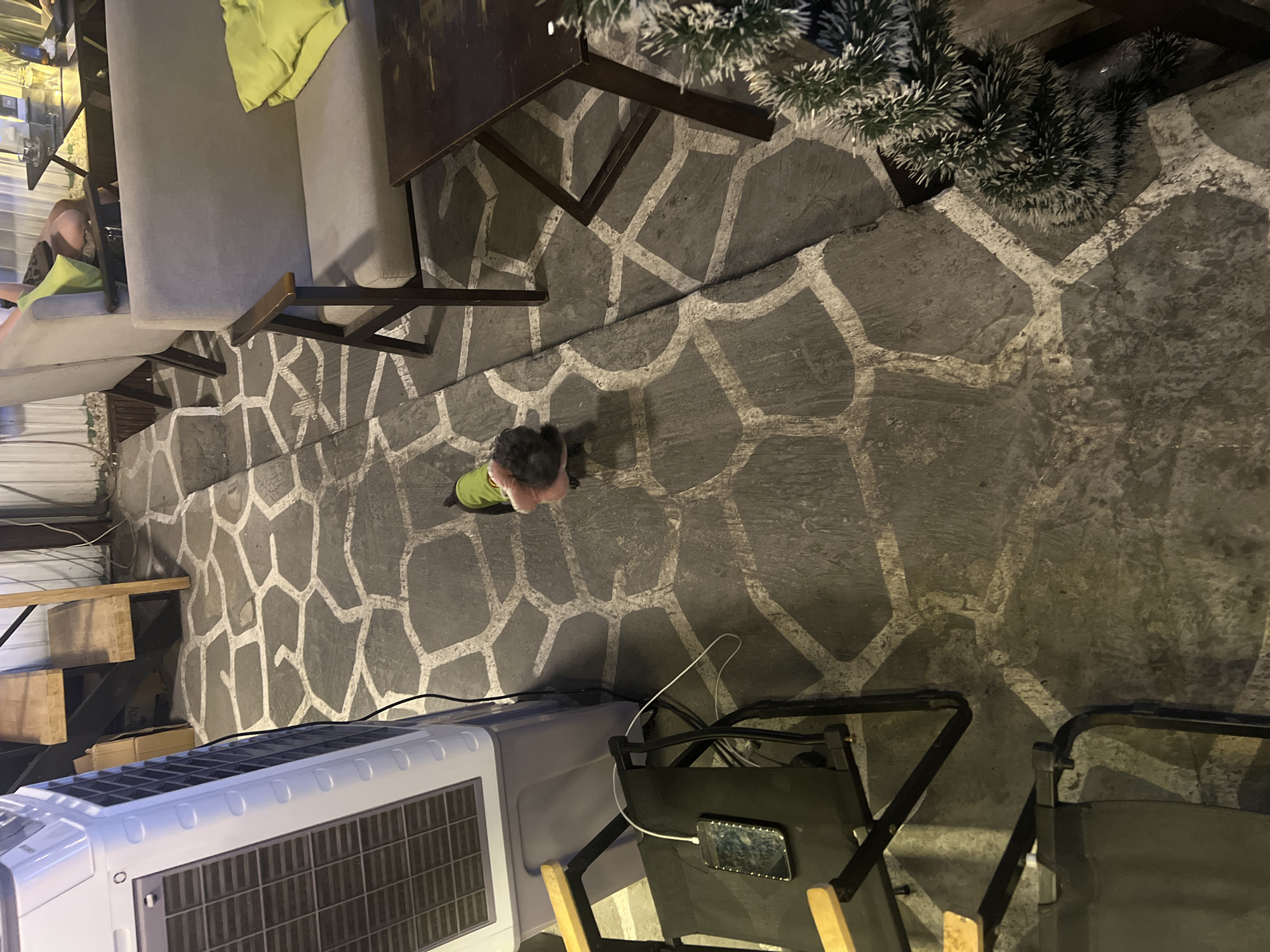 This image, captured from an overhead perspective, depicts a relaxed indoor or covered outdoor area, likely a cafe or lounge in Đà Nẵng, Vietnam, during the evening or night.
The dominant feature is the dark grey patterned floor, composed of irregularly shaped stones or concrete sections outlined by striking white grout lines. In the center of the frame, a small child with dark hair is seen from above, standing on this patterned floor. The child wears a light green jacket over a pink garment.
To the left, two grey armchairs or sofas with light green cushions provide seating, with a person's legs partially visible on one of them. In the upper right section, a dark, possibly wooden, table with metal legs is situated near an artificial evergreen plant, resembling a small, frosted pine or fir tree, adding a decorative touch.
The lower left corner shows a white portable air conditioner or cooler on the floor. Next to it are two dark fabric folding chairs with wooden armrests. A smartphone is resting on one of these chairs, visibly connected to a white charging cable.
The scene is lit by artificial light, suggesting it is nighttime or late evening. The presence of the air conditioner hints at a warm climate. No discernible text is visible in the image. The overall impression is of a casual and comfortable commercial or social setting.