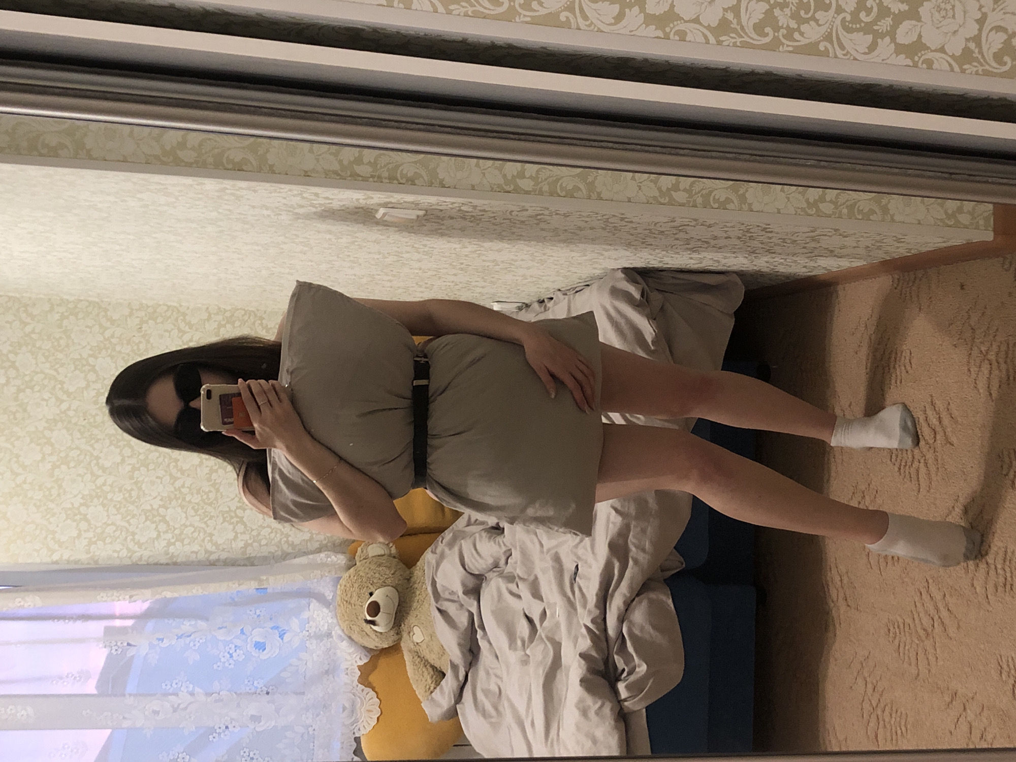 A young woman is captured in a mirror selfie indoors, participating in what appears to be a "pillow challenge." She stands, with her body angled, against a bed or sofa, wearing a large gray pillow cinched at the waist with a dark belt, resembling a dress. Her long dark hair frames a face obscured by sunglasses and a light-colored smartphone she holds up to take the photo. Her bare legs are visible, ending in white ankle socks.
The scene is reflected in a large mirror, likely part of a wardrobe. Behind the reflection of the woman, a light brown teddy bear with a heart on its chest sits on a yellow pillow next to crumpled gray bedding on a dark blue bed frame. The walls feature light-colored patterned wallpaper, and a window with white sheer floral curtains reflects soft, diffused light, suggesting either morning or late afternoon. The floor is covered with a light brown, textured carpet. The overall impression is a casual, relaxed atmosphere within a private residence in Noviy, Russia. No visible text is present in the image.