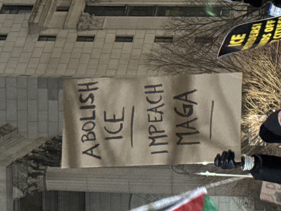A tan protest sign reads "Abolish ICE" and "Impeach MAGA" in vertical, block lettering with an underscore separating each phrase. A partially visible sign reading "ICE Murderers! Hands Off Cities!" is at the top right of the image. The sign is held at right by a person in a black coat and black gloves. A large stone building and bare trees are in the background. A partial view of a red and green flag is at the bottom of the image.