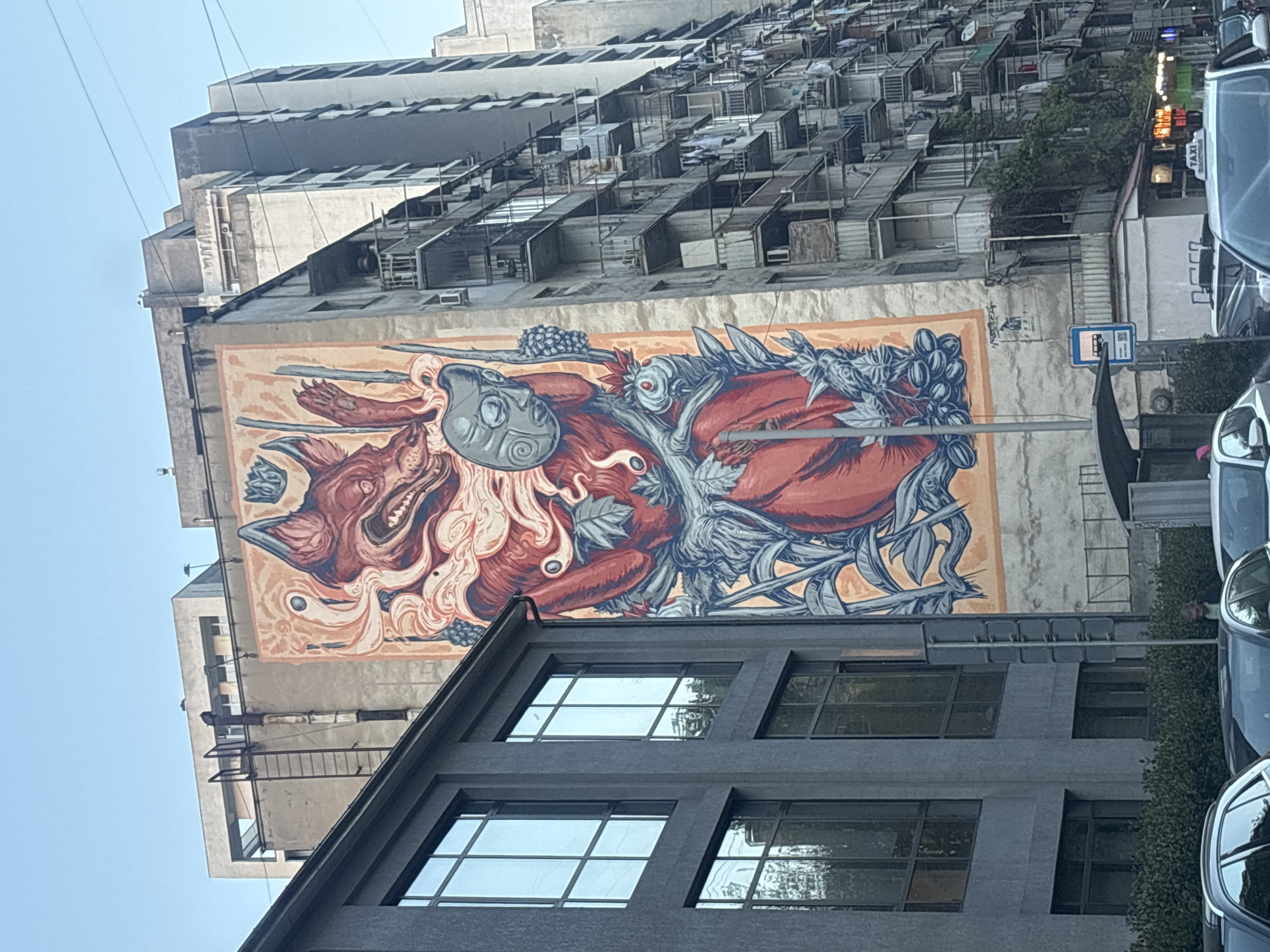 The image displays an urban scene in Tbilisi, Georgia, centered around a large, vibrant mural painted on the side of a tall, light-colored concrete building. The mural features two main figures in a stylized, dynamic composition. One figure appears to be a humanoid wearing a mask, while the other is a snarling, canine-like creature. Both are intricately intertwined with leafy branches and clusters of berries, rendered in dominant shades of deep red, blue-gray, and orange-brown.
The building hosting the mural is a high-rise structure, appearing somewhat aged, with numerous small, box-like extensions or makeshift balconies protruding from its upper floors. In the foreground, a modern, dark grey building with large rectangular windows occupies the lower portion of the frame, providing a visual contrast to the older structure and the artwork.
The scene is set during the daytime, under a clear, bright sky, indicated by the even lighting. Parked vehicles are visible on the right side of the image, partially obscured by hedges and the foreground building. Visible text includes a blue and white sign depicting a bus icon, likely indicating a bus stop, and a partial "TAXI" sign on top of one of the cars. The overall impression is a striking blend of street art and urban architecture.