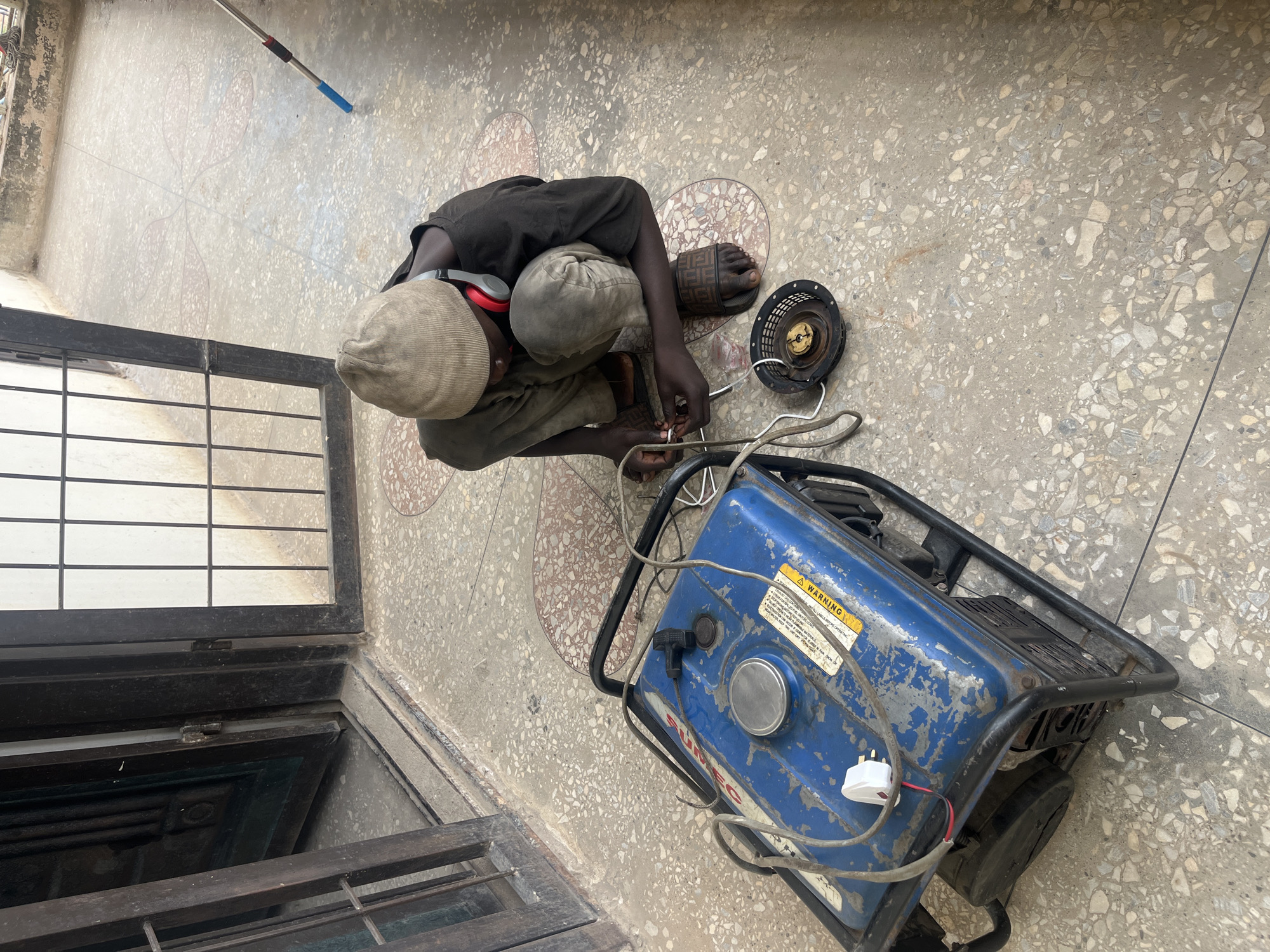 In Gura Top, Nigeria, a young boy wearing a beanie and headphones is crouched on a terrazzo-tiled outdoor patio, working on a blue generator. The patio has a decorative pattern of pink and beige tiles. Adjacent to the generator, a black starter recoil assembly is on the ground. The boy is manipulating wires connected to the generator. Behind him, a dark metal-framed doorway with a glass pane and bars stands open. Sunlight illuminates the scene, casting shadows on the ground.