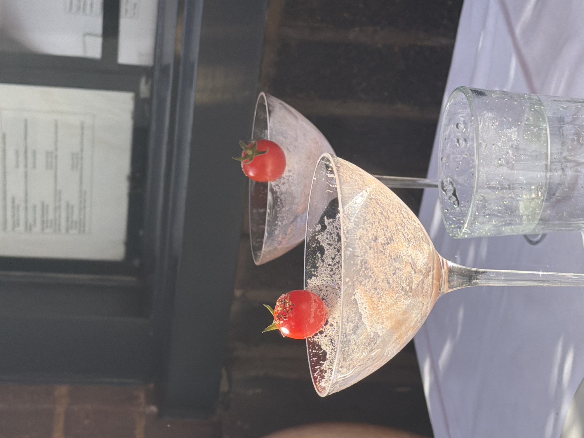 The image shows two martini glasses with cherry tomatoes as garnishes, positioned next to an empty water glass. The glasses are set against a backdrop of a white tablecloth and a window or door frame with a menu or notice displayed. The location is the city of London, United Kingdom.