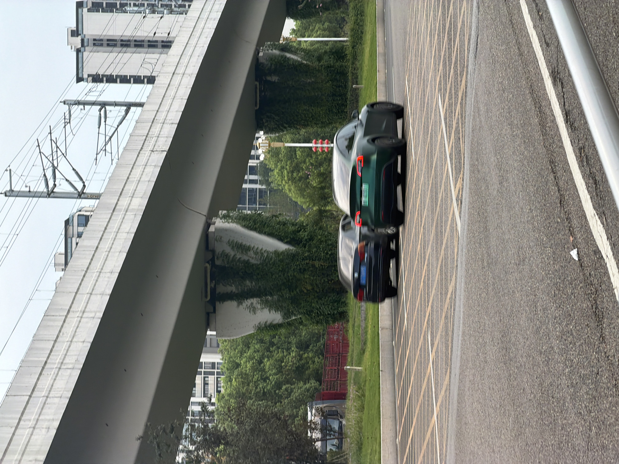 A green Tesla Model 3 and a dark sedan are captured in motion on an urban street in Jiangjin, China. The vehicles are positioned in the foreground, with the Tesla in the right lane and the sedan in the left lane, both facing away from the viewer. In the background, a multi-story concrete overpass dominates the scene, with power lines and supporting pillars visible. Lush green trees and modern buildings are interspersed between the overpass and the sky. The street is marked with yellow and white lines, indicating traffic lanes and a possible intersection or parking area. The overall scene suggests a typical day in a developed urban environment with active transportation infrastructure.